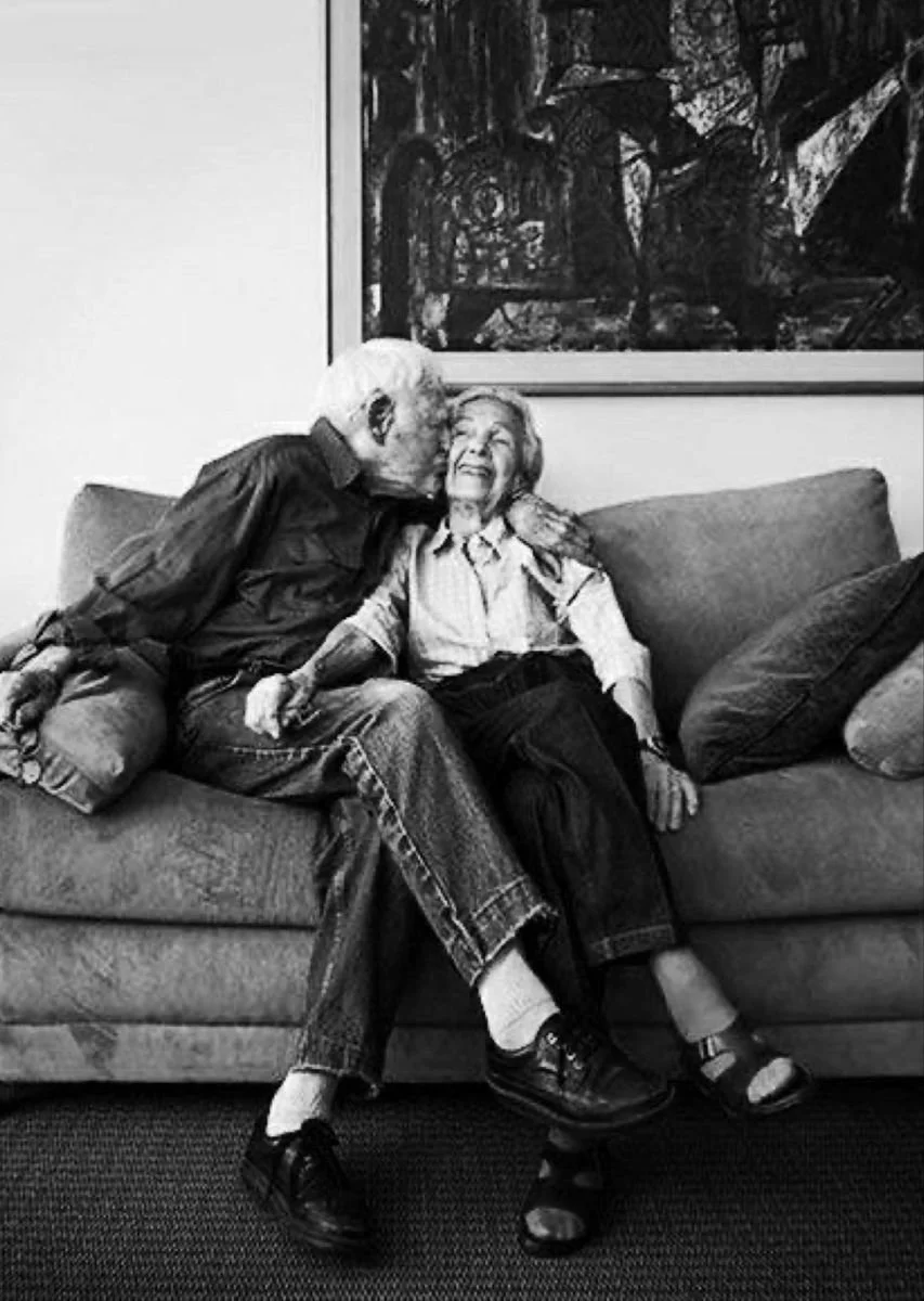 An elderly man is kissing an elderly woman on the cheek while they sit together on a couch. The woman is smiling happily, and there is a large abstract painting hanging on the wall behind them.