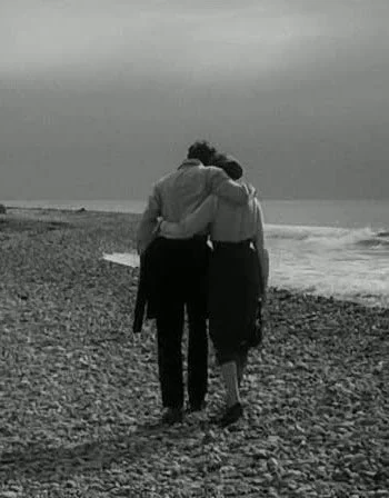 Two people walking arm in arm on a pebble beach, facing the ocean under a cloudy sky.