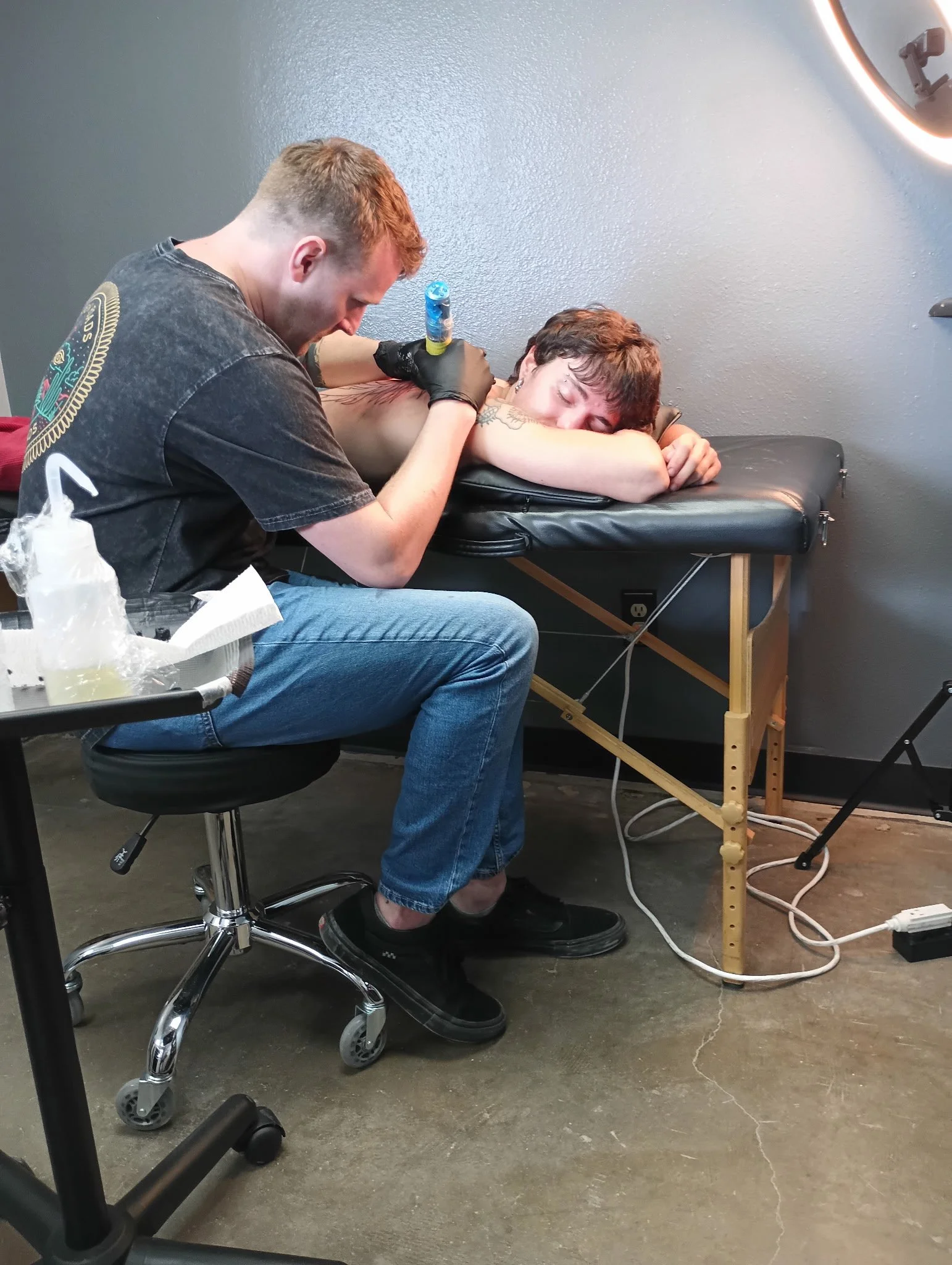 Tattoo artist tattooing a full back-piece while the client lies on a black massage table in a tattoo studio.