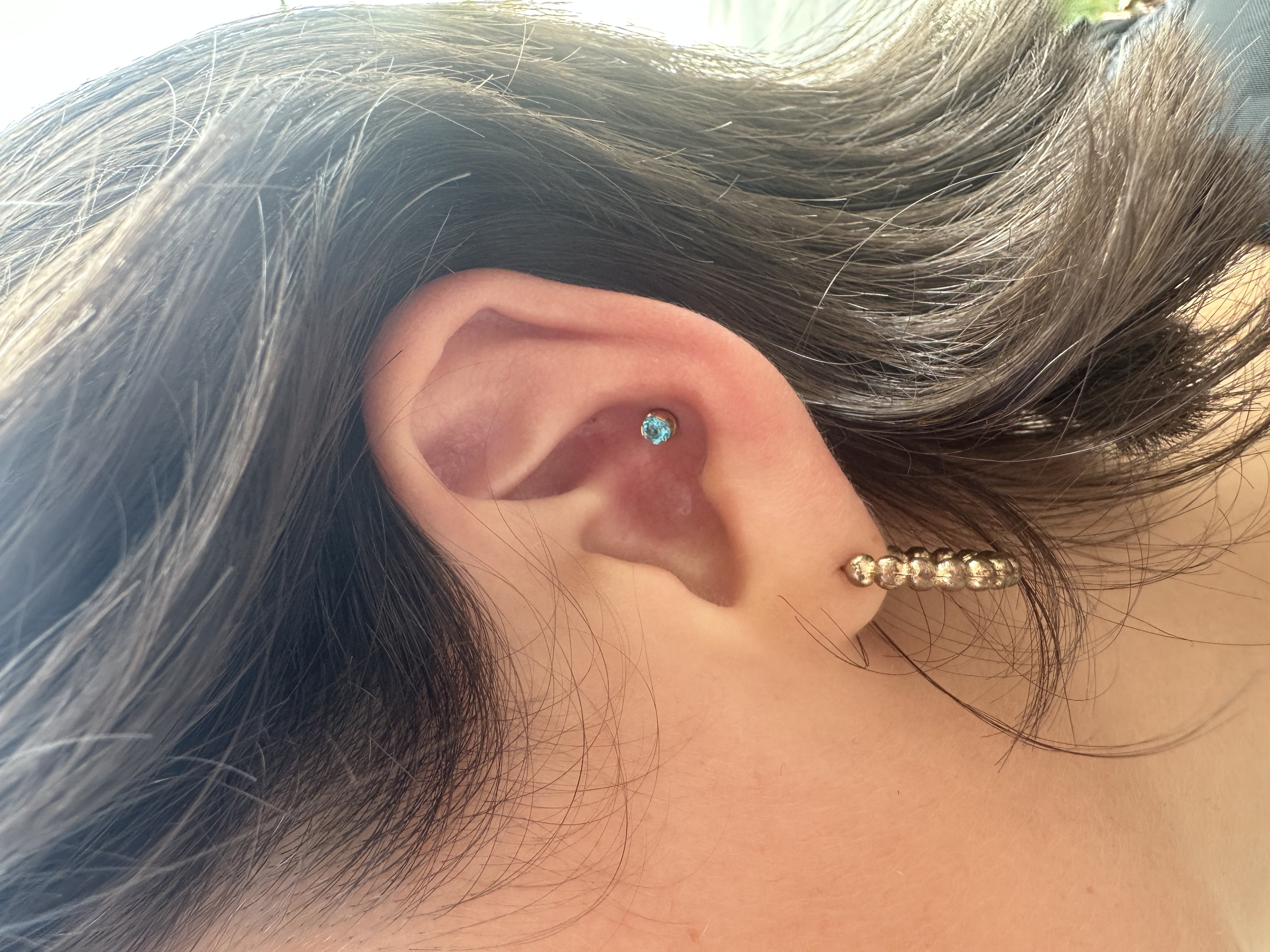 Close-up of a person's ear with three piercings: a blue gemstone in the helix, a gold ball in the tragus, and a row of small gold balls on the earlobe.