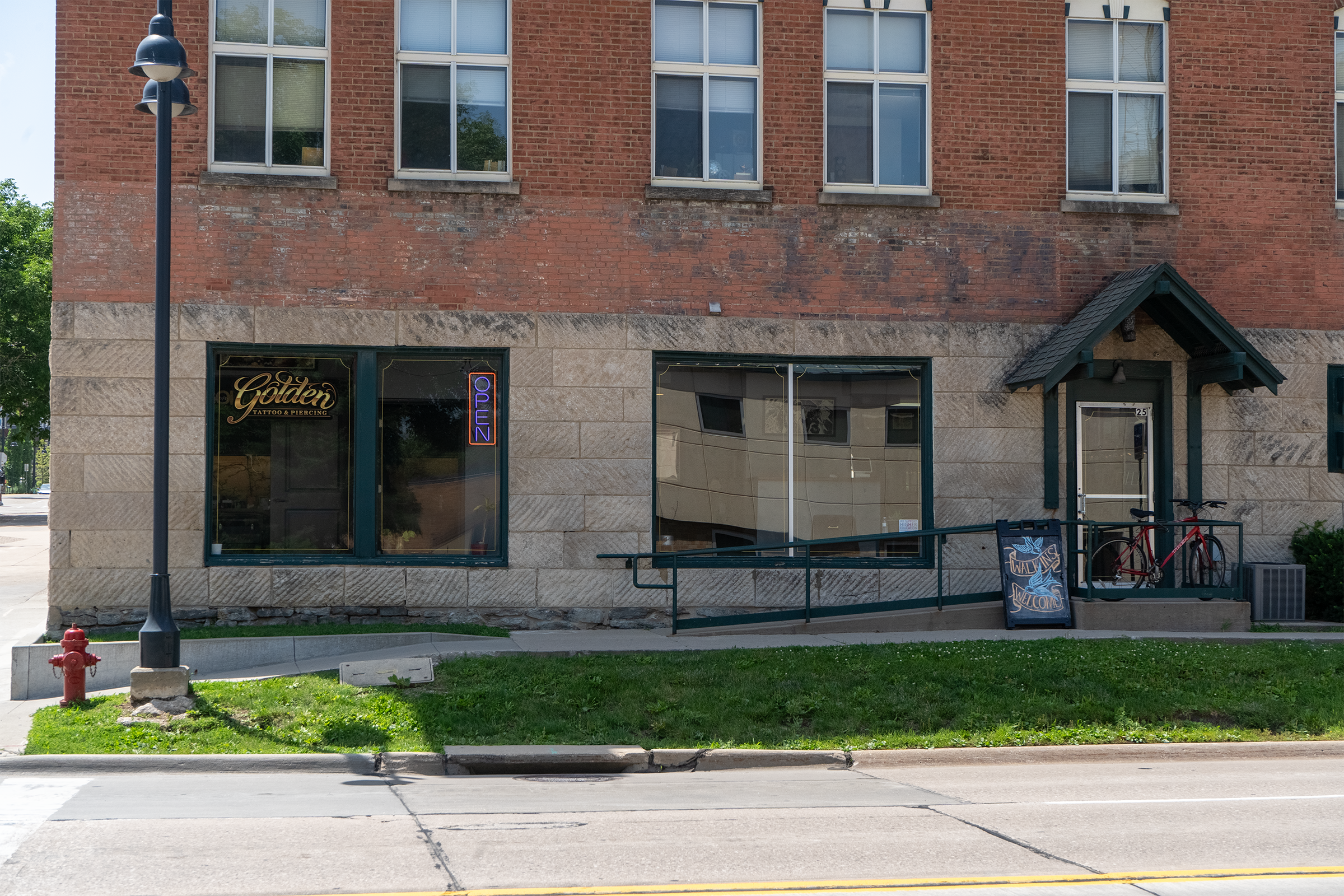 The image shows the exterior of a brick building with large windows and a storefront on the ground level. There is a black sign that reads "Golden Tattoo & Piercing" next to a neon "Open" sign in the window. A small outdoor area with bicycles, a sidewalk, some greenery, and a parked red fire hydrant is visible in the foreground. A street runs parallel to the building.