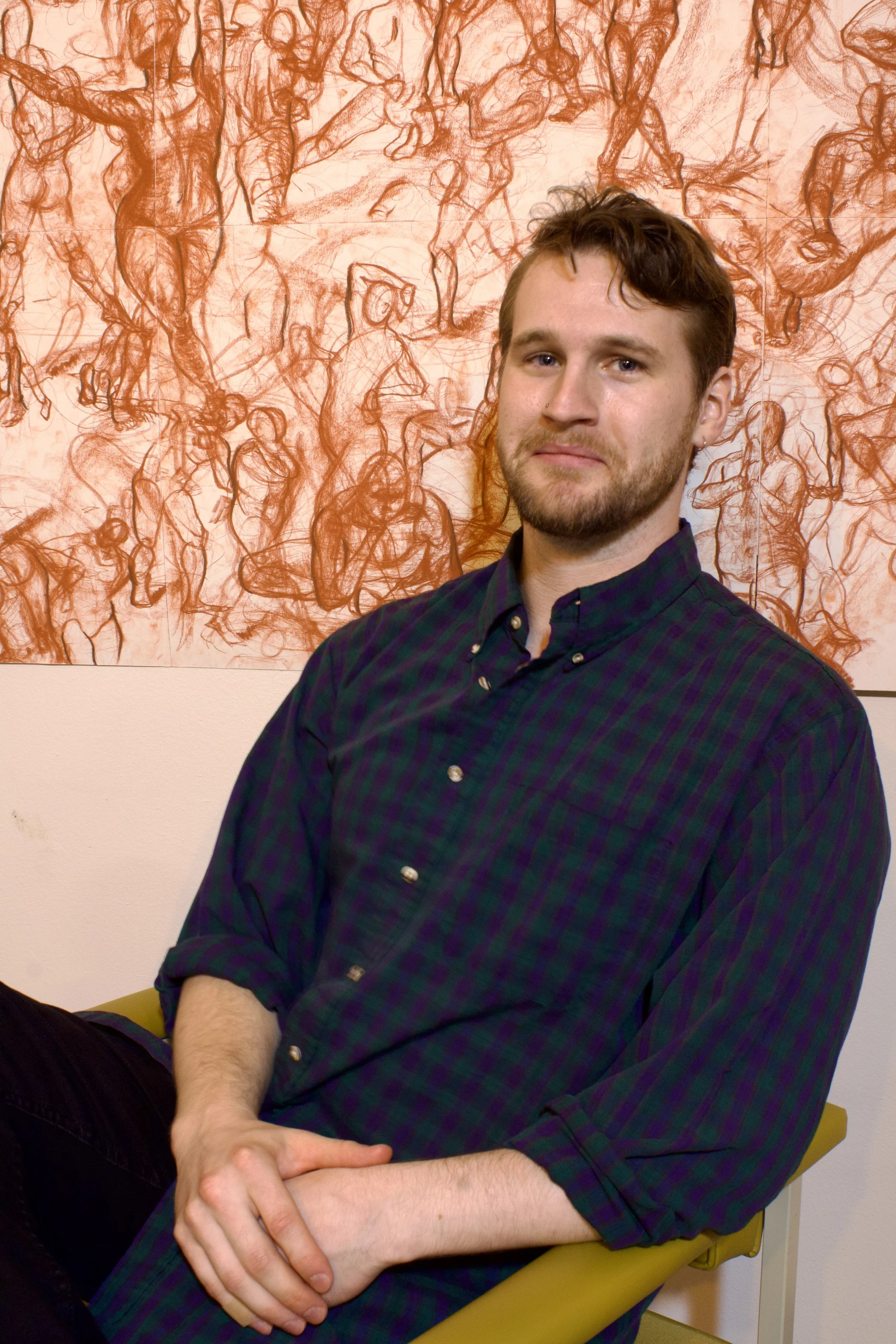 Portrait of a man with a beard and wavy hair sitting on a yellow chair, wearing a dark plaid shirt with rolled-up sleeves, with an abstract red and white drawing of various human figures in the background pastel pencils. Art School.