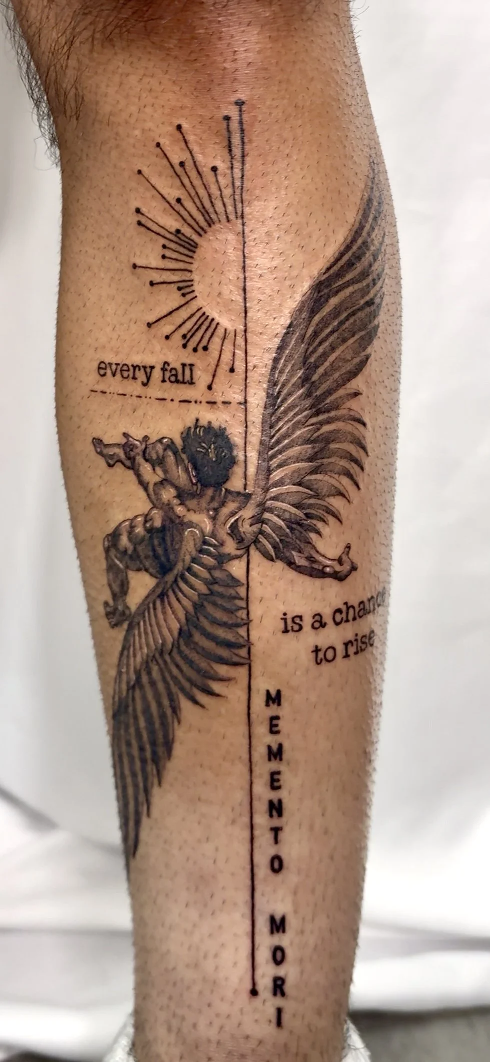Tattoo of a winged cherub or angel in a dynamic pose, with a line dividing the tattoo into two parts. The left side features a sun with rays, and the right side contains a quote that reads 'every fall is a chance to rise.' The vertical line has the w