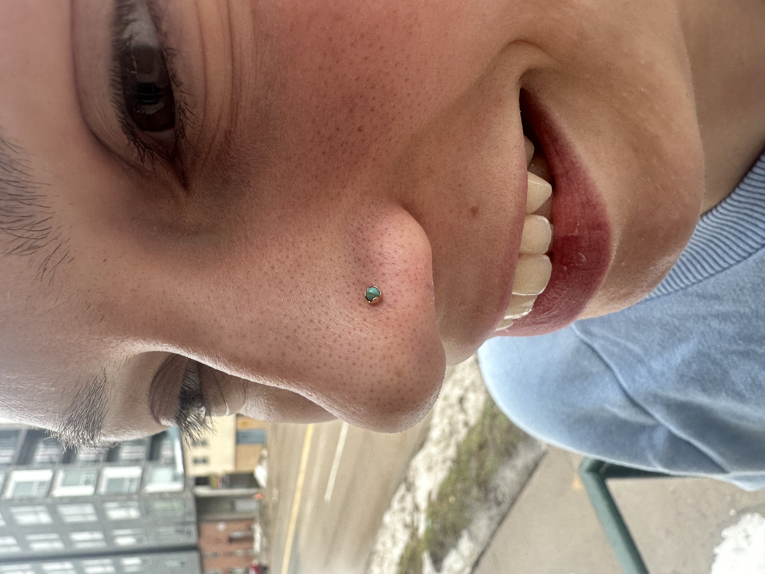 Close-up of a person's face showing a nose piercing with a small turquoise stud, smiling with teeth visible, wearing a light blue shirt, outdoor urban background.