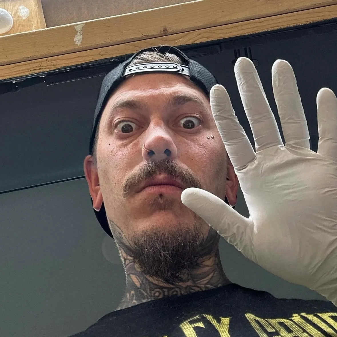 A man with a mustache and a tattooed neck is looking into the camera, wearing a baseball cap backwards and a white medical glove, holding his hand close to the camera. Experienced Piercer