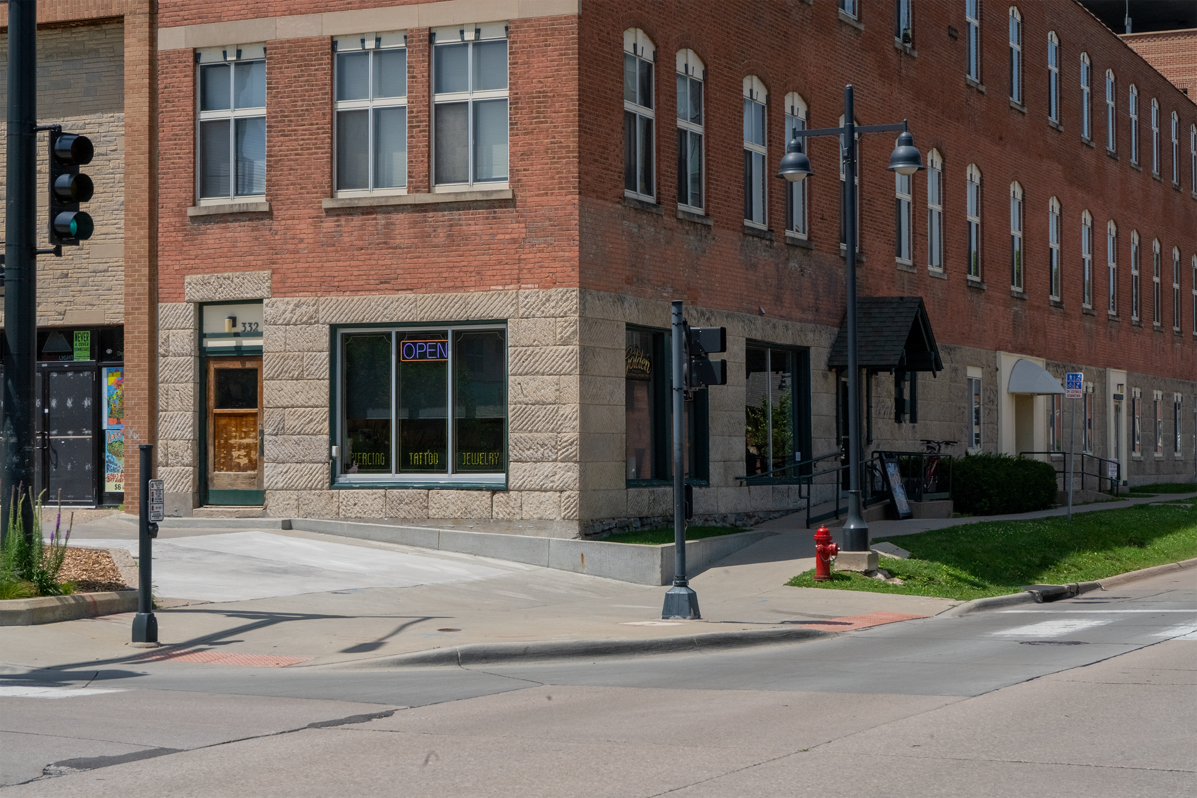 Street view of a brick building corner with a storefront that has a neon 'OPEN' sign, and neighboring shops, including a tattoo and jewelry shop, at 332, with traffic lights, street lamps, and a fire hydrant.