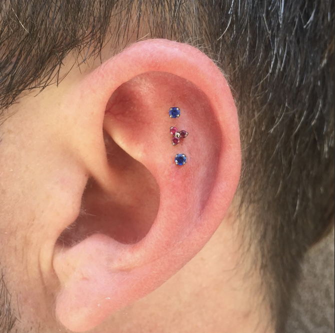 Close-up of a person's ear with three small stud piercings, two with blue gemstones and one with a pink gemstone, arranged vertically.