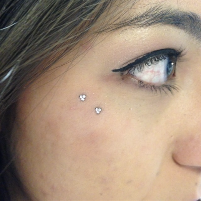 Close-up of a person's face showing eyes and cheek with a small sparkle tattoo near the eye, makeup with winged eyeliner, and a small cluster of rhinestones on the cheek.