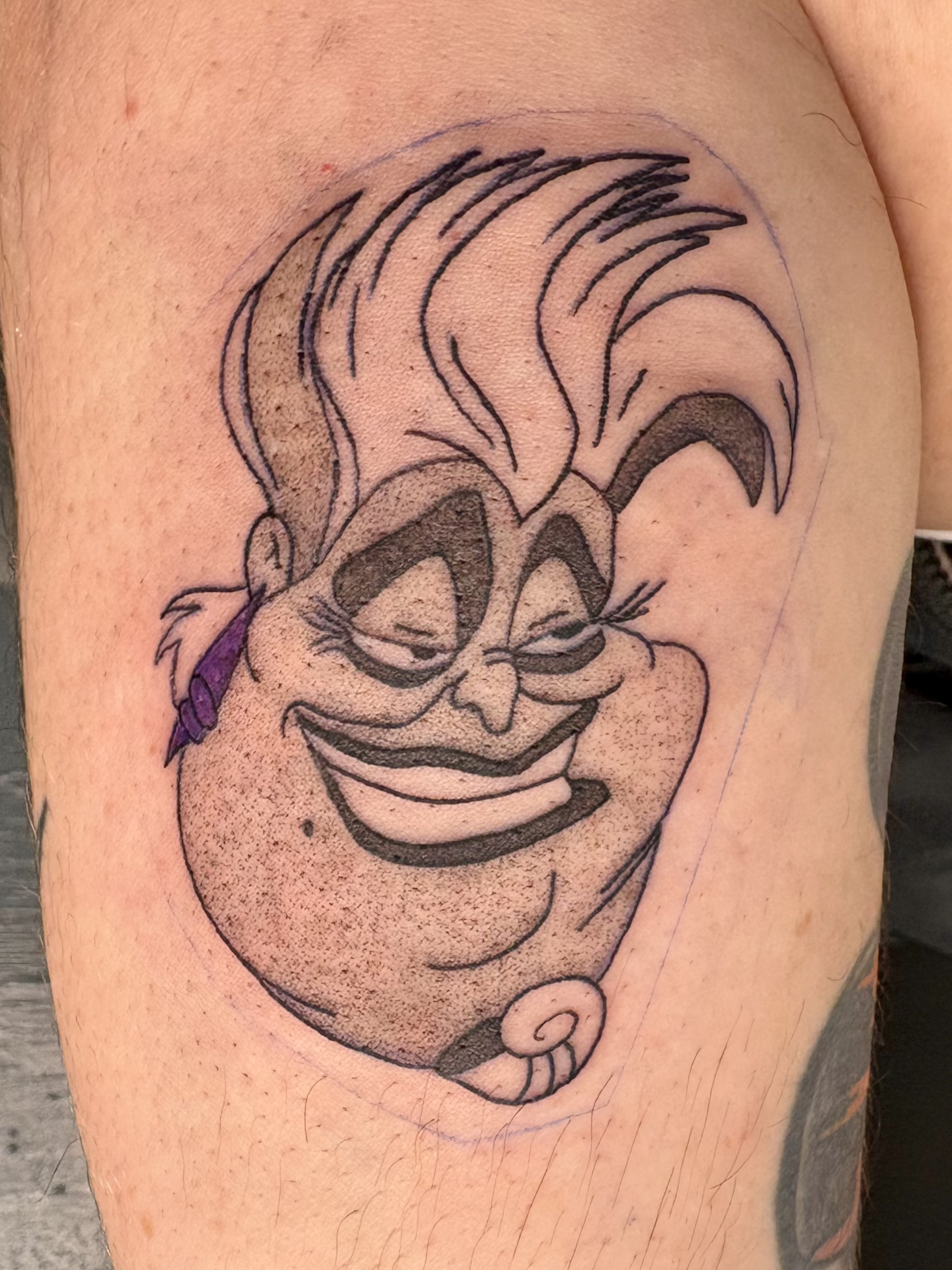 Tattoo of a grinning cartoon clown face with exaggerated features, spiky hair, and a small purple bow on the side. Stipple Shading.
