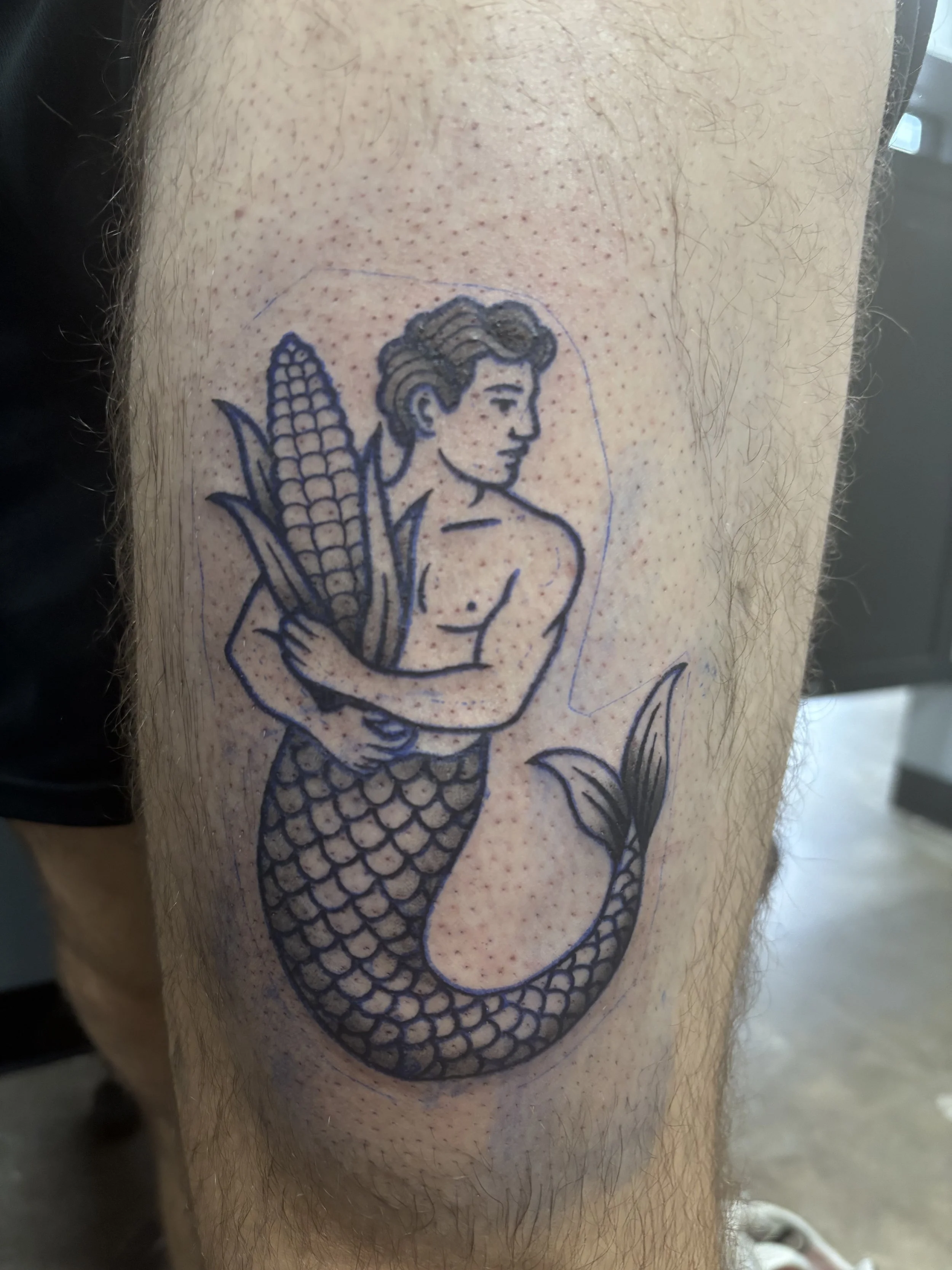 American Traditional Tattoo of a mermaid with a human upper body and fish tail, holding ears of corn.