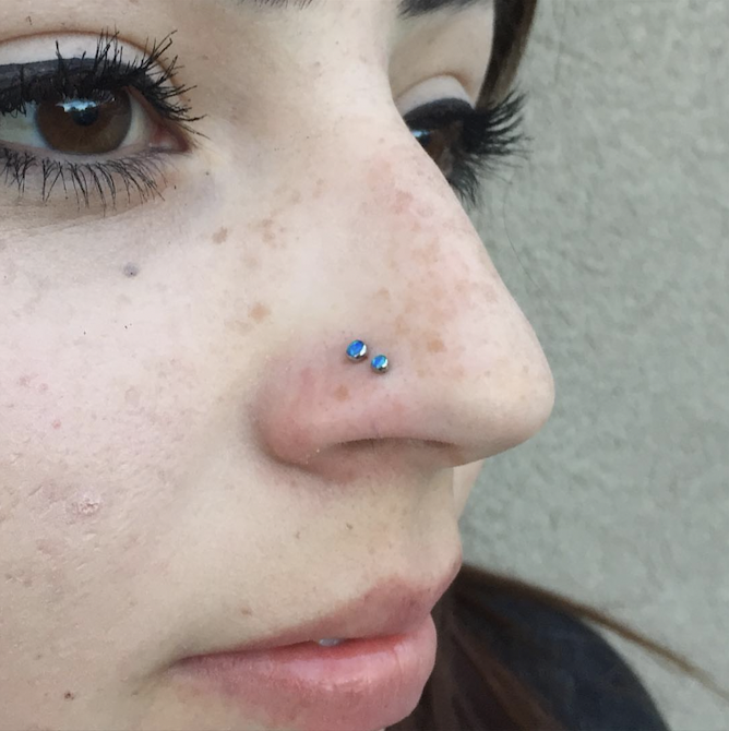 Close-up of a person's face showing their nose with two small blue gemstone studs and a side of their eye with makeup, against a neutral background.