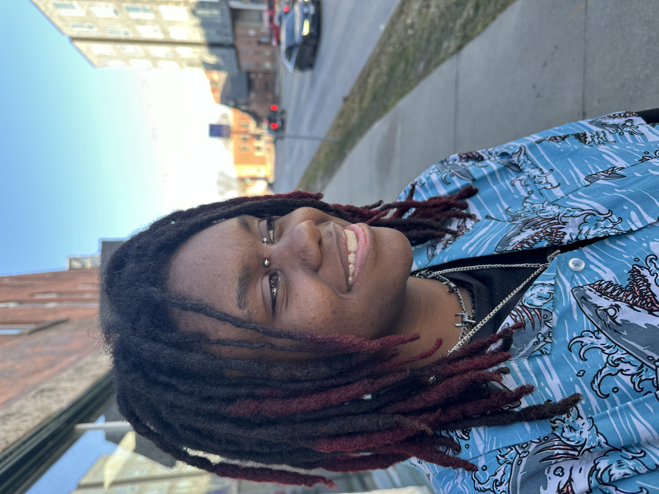 A young man with dreadlocks and facial piercings smiling outdoors on a city street, wearing a patterned blue jacket with ocean wave design.