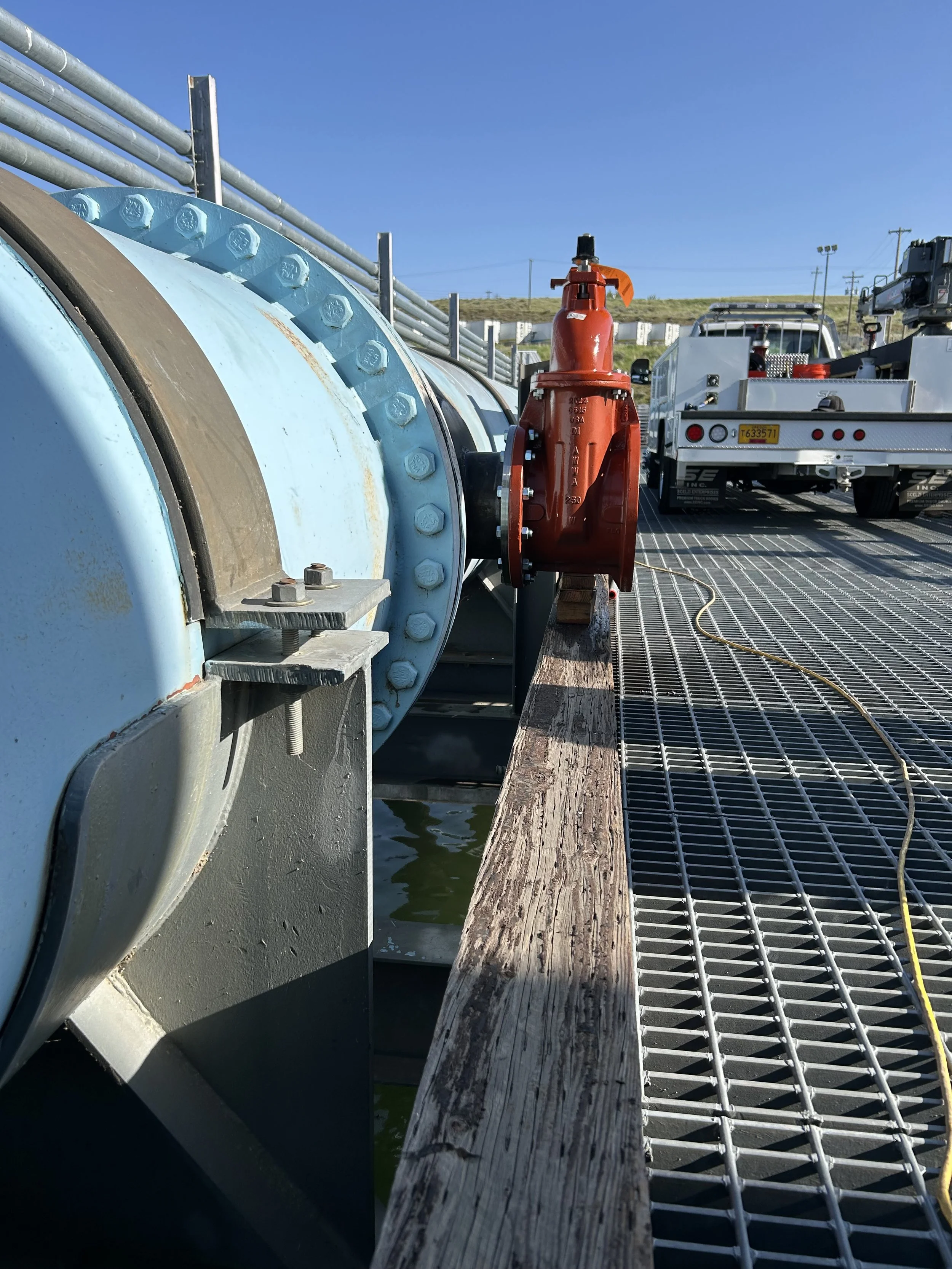 Close-up of a large blue industrial pipe with bolts, next to a red fire hydrant, on a metal grate dock with a utility truck in the background under a clear blue sky.