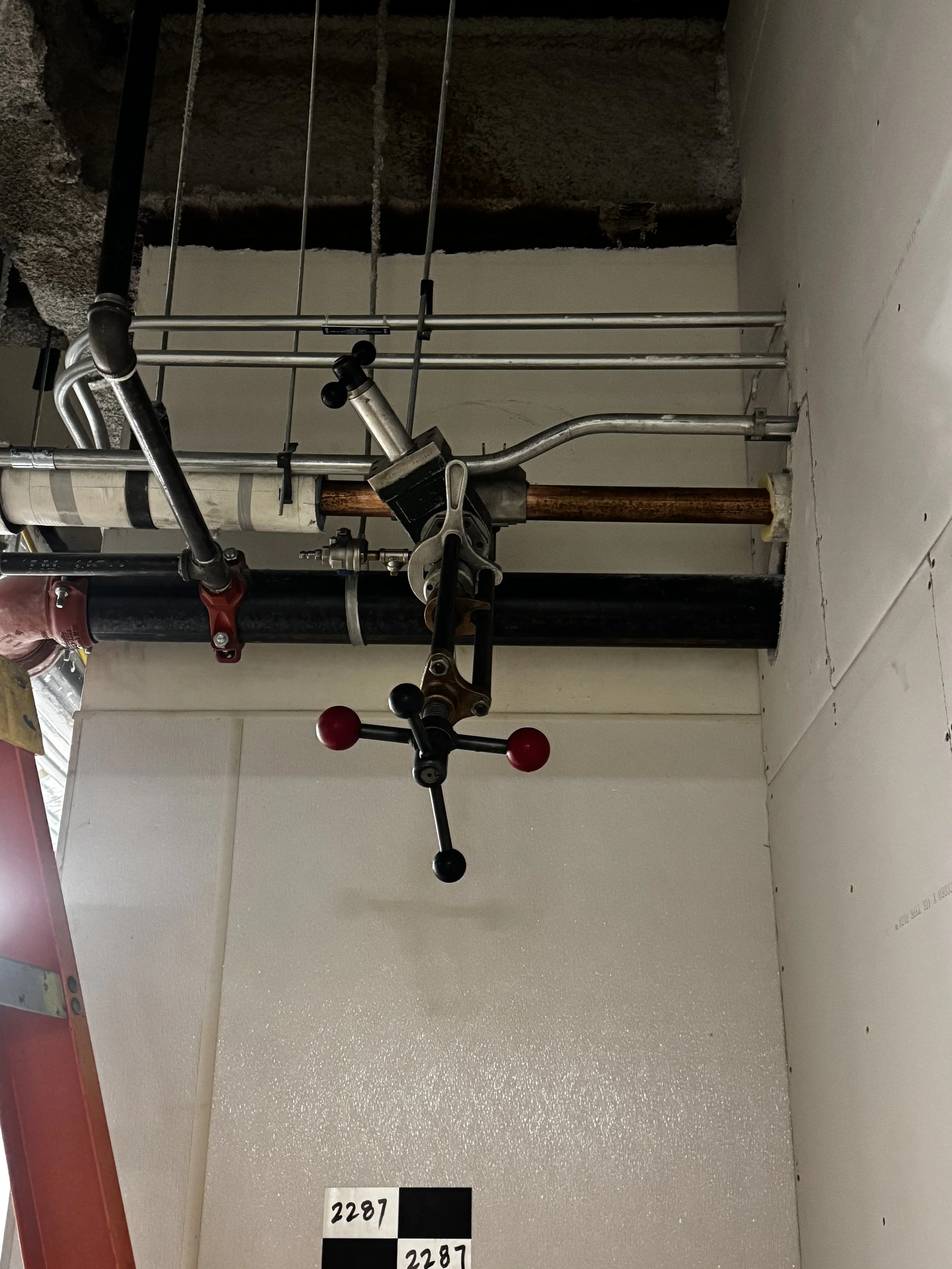 Construction site with pipes, a pipe clamp, and a level tool installed on a ceiling, with drywall and unfinished wall area visible.