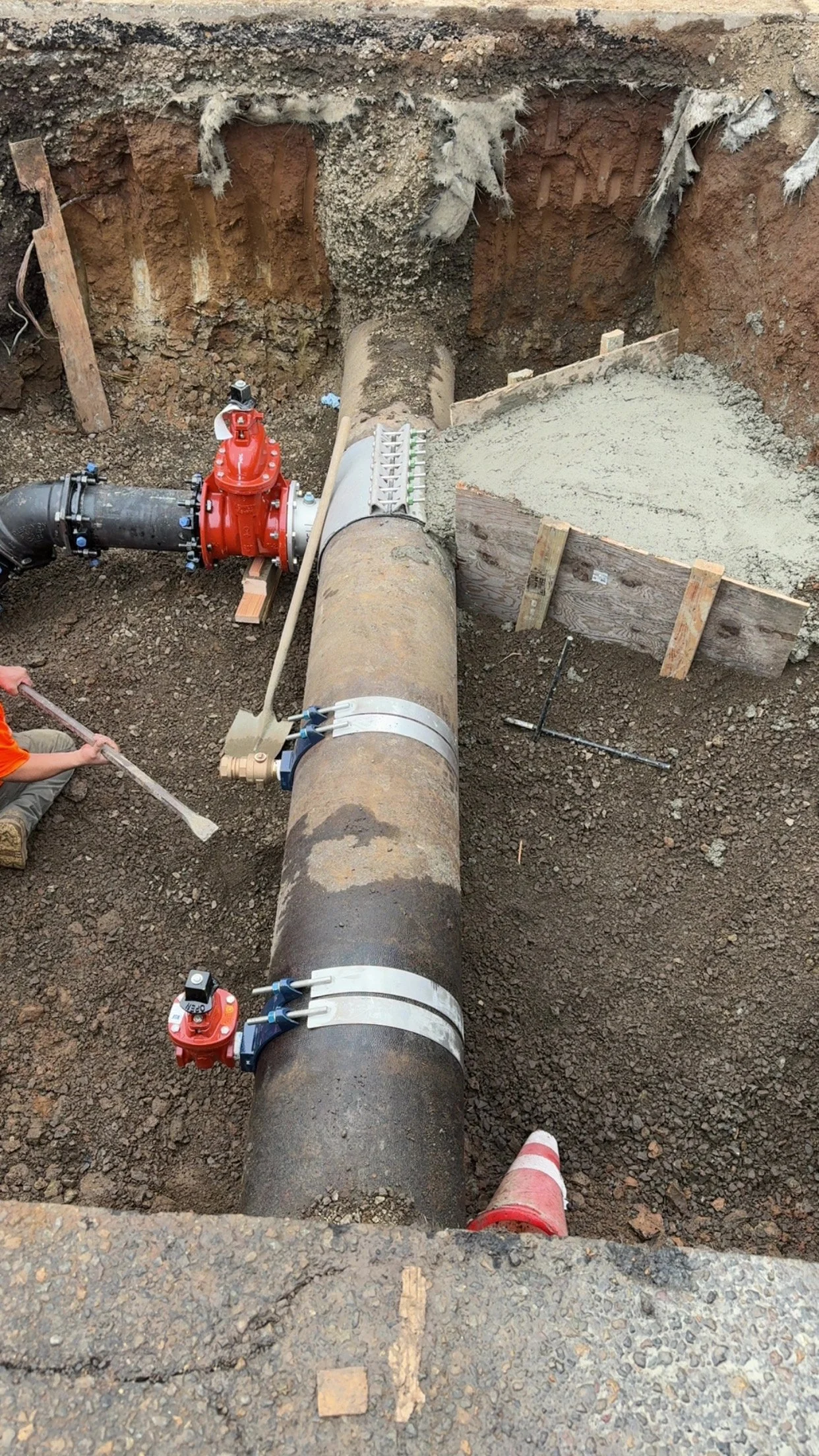 Construction worker working on underground pipe installation, with large pipe, valves, and concrete footing in excavation site.
