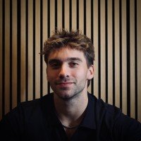Close-up of a smiling woman with curly hair, wearing glasses, hoop earrings, and layered necklaces, in a brightly lit indoor space.