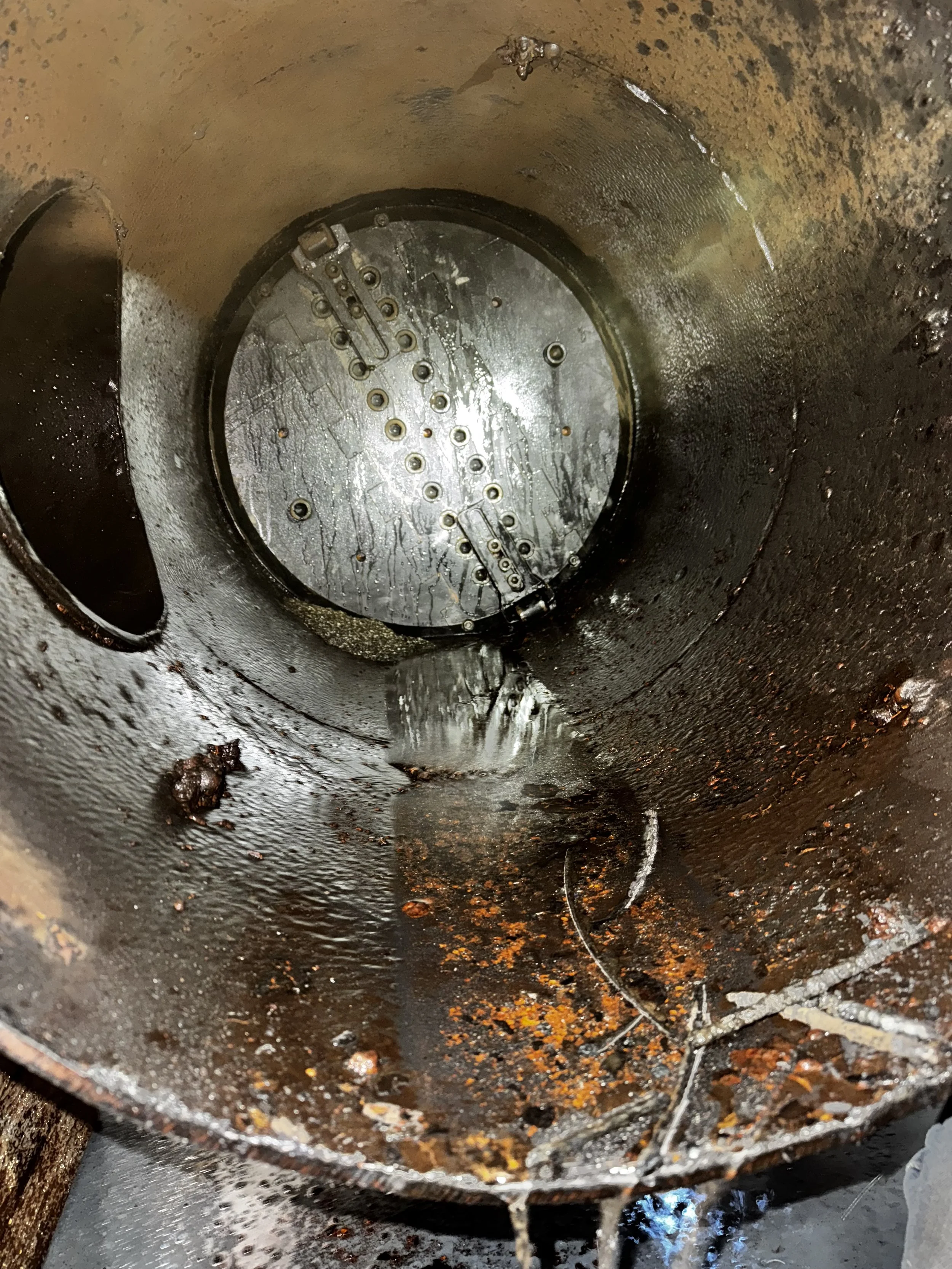 Inside oil or fuel storage tank with rust, water, and debris, showing a metal inspection or access plate at the top.