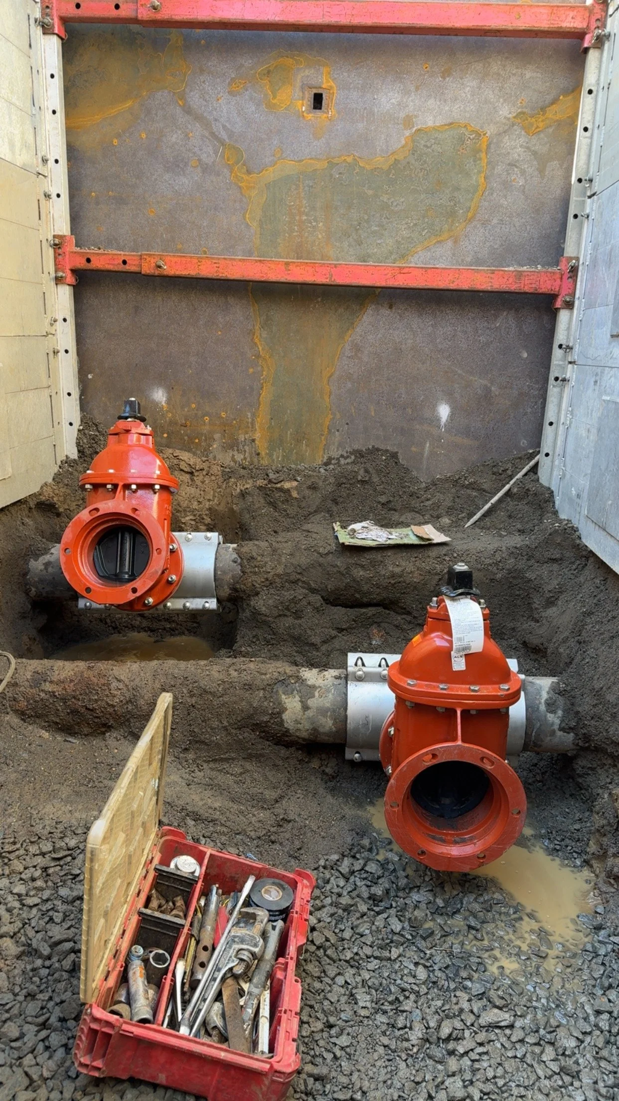 Construction site with two red water control valves connected to large gray pipes, dirt and gravel on the ground, and tools in a red toolbox nearby.