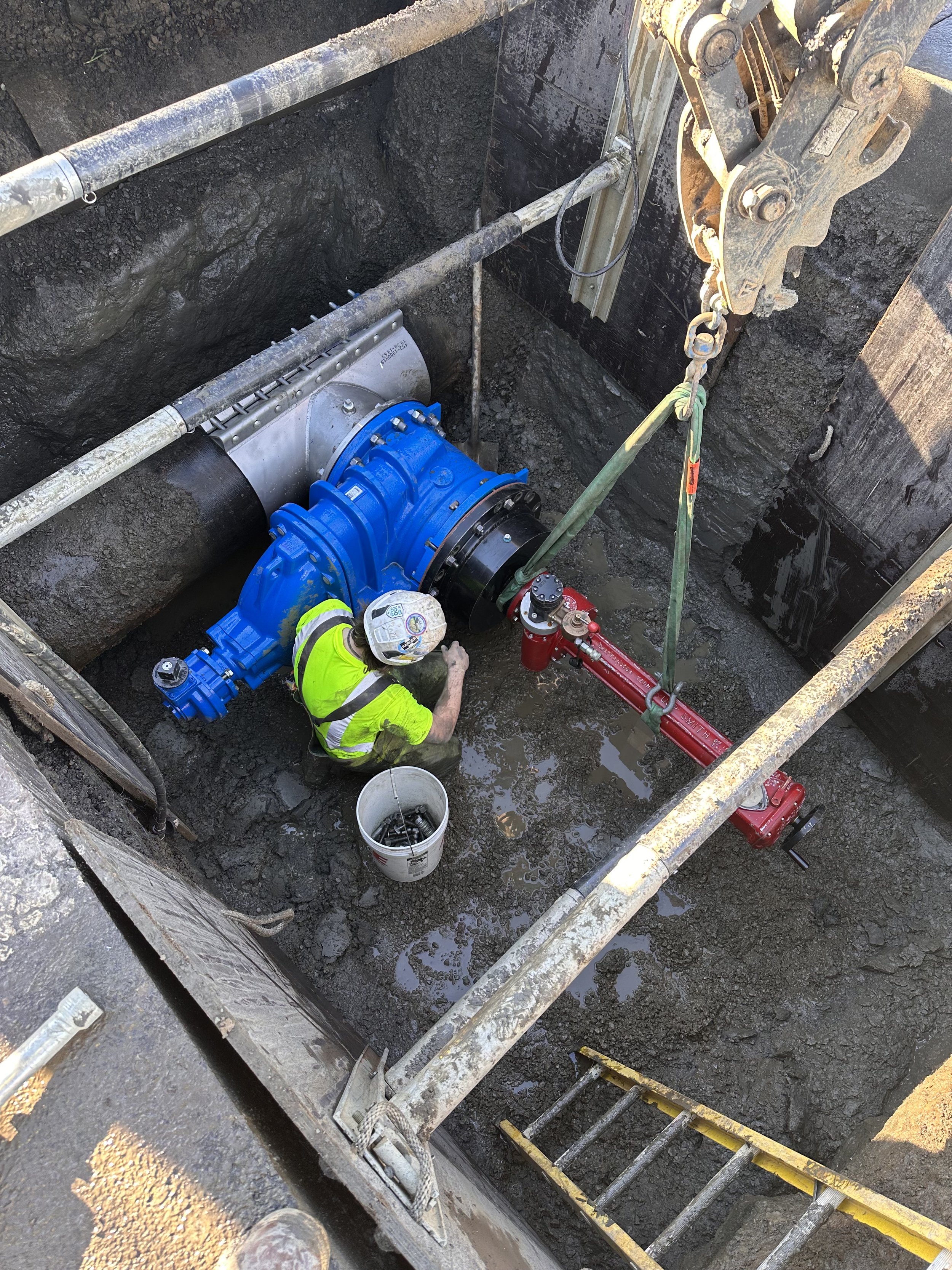 Residential or commercial construction worker in a safety helmet and reflective vest working on large blue and red machinery in a construction trench, with scaffolding and muddy ground surrounding the work site.