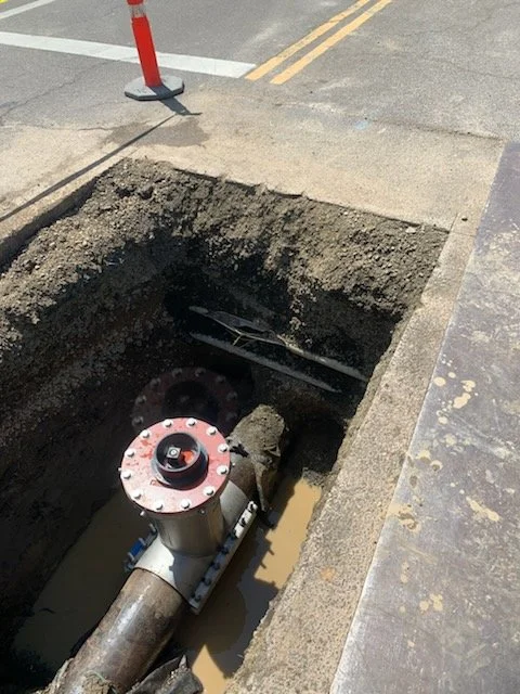 Open street road excavation exposing underground water or sewer pipe with valve insertion, halted traffic cone nearby.
