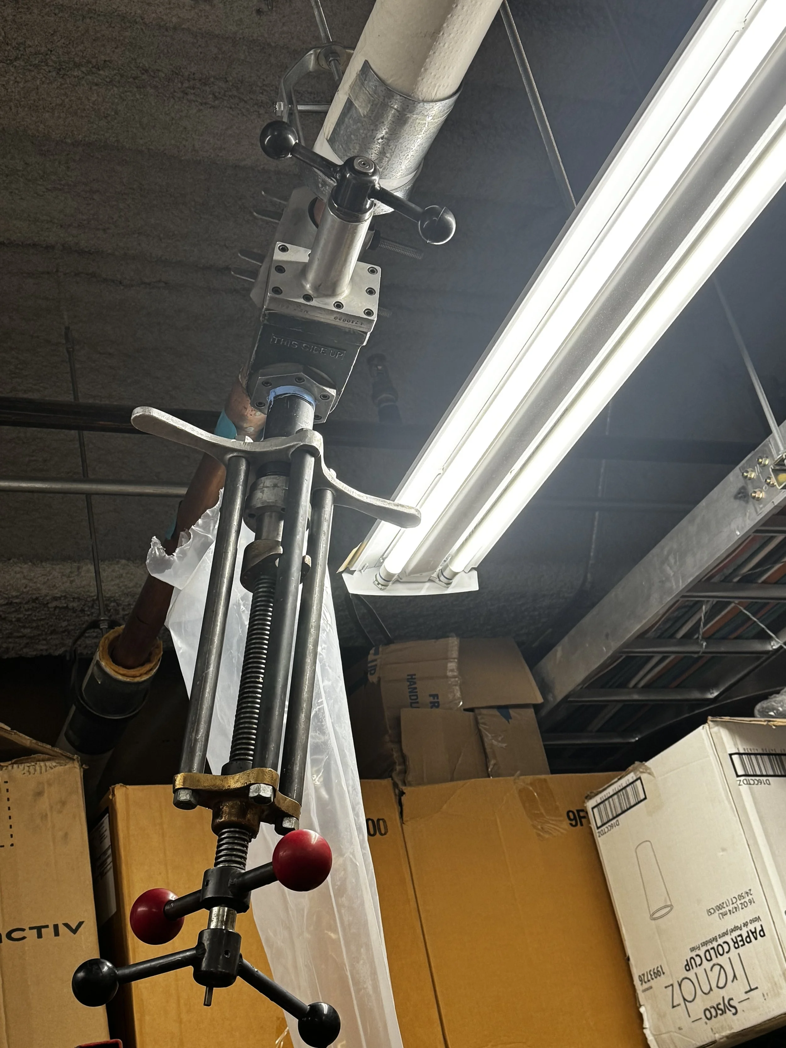 Industrial piping and fixture in a warehouse setting with fluorescent lights and cardboard boxes in the background.