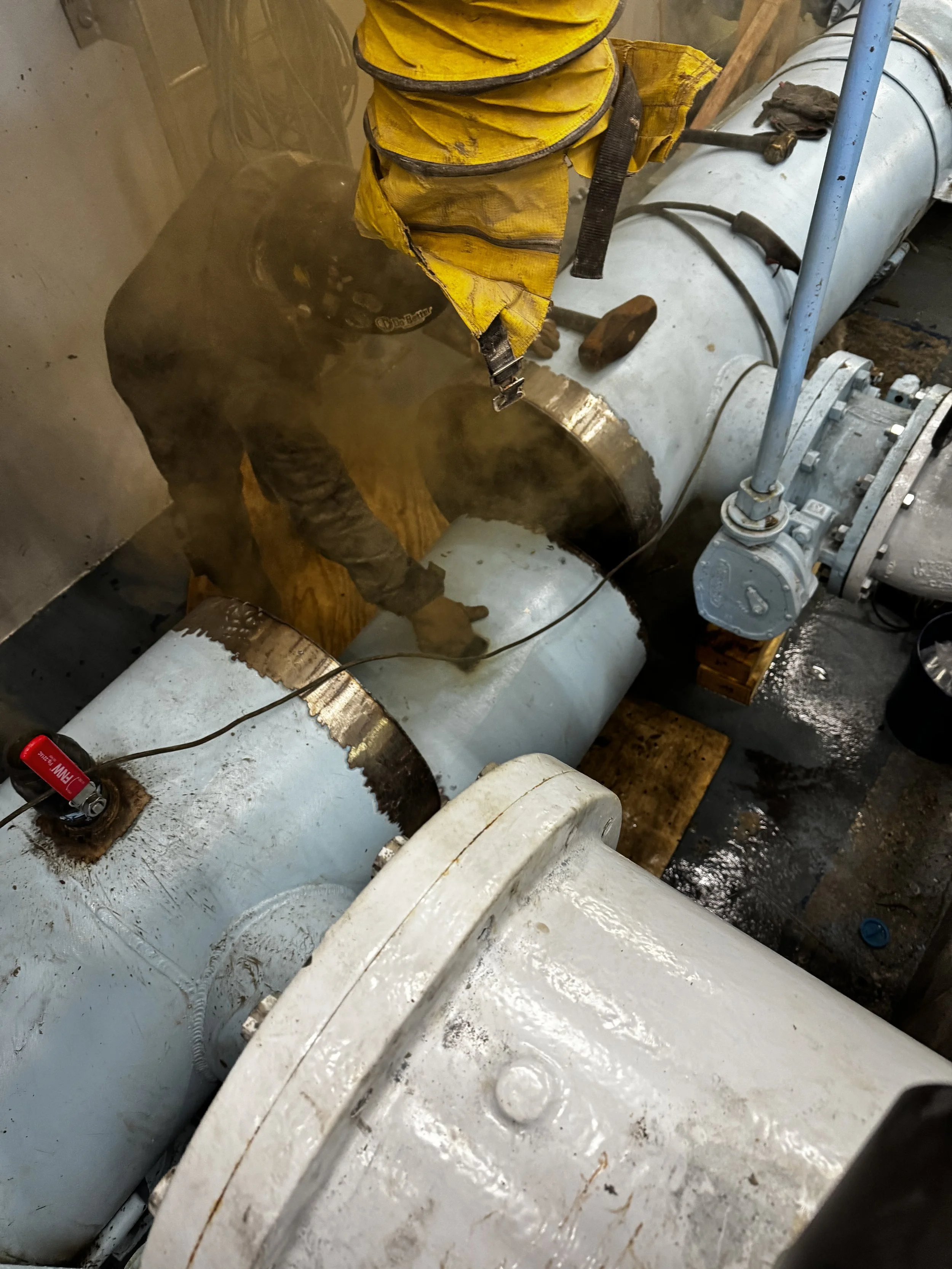 A worker wearing a yellow safety vest and gloves is welding a pipe in an industrial setting, with sparks and smoke around the welding area.
