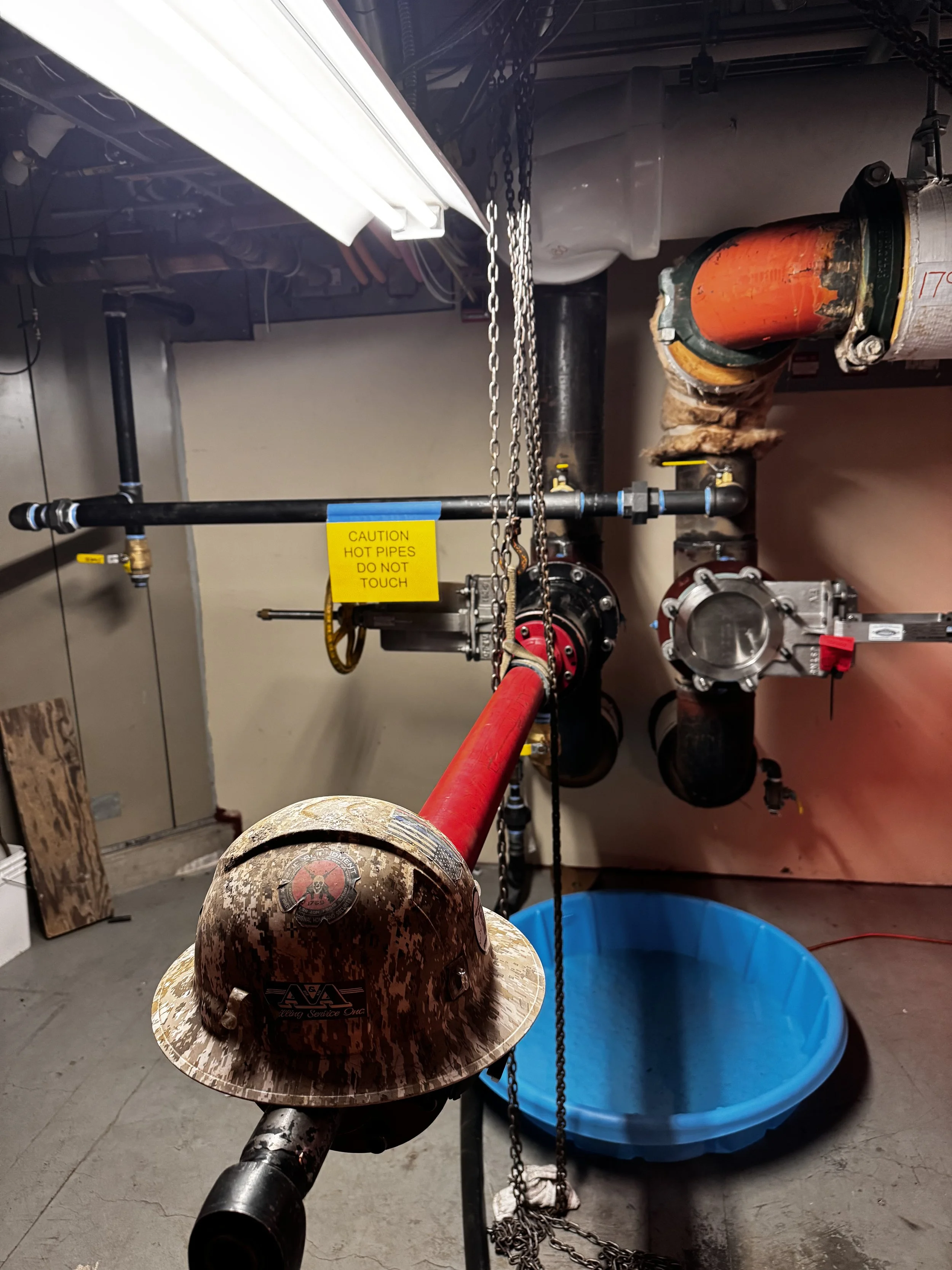 Image of industrial pipes and equipment in a utility room, including a red pipe with a warning sign, and a construction helmet placed on one of the pipes, with a blue basin on the floor nearby.