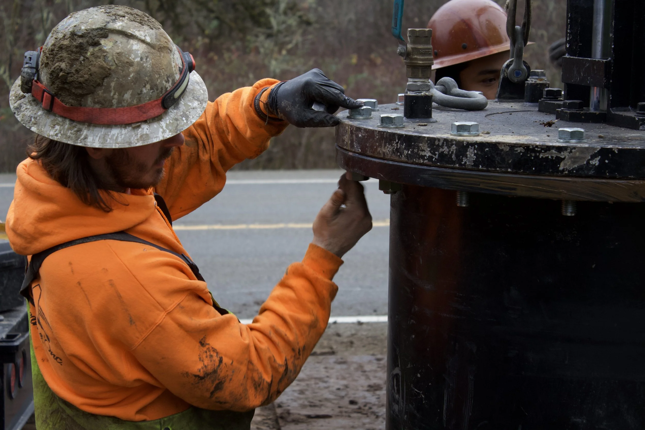 Worker inspecting or working on a large pipe or industrial equipment on the side of a road, wearing safety gear including orange jacket and helmet.