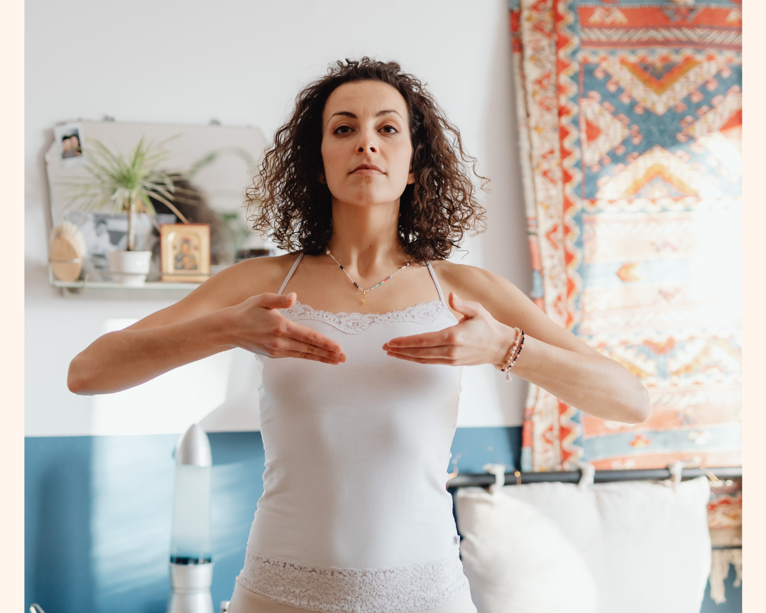 A woman with curly hair standing indoors, holding her hands in front of her chest in a meditative or calming gesture, with a serious or focused expression.