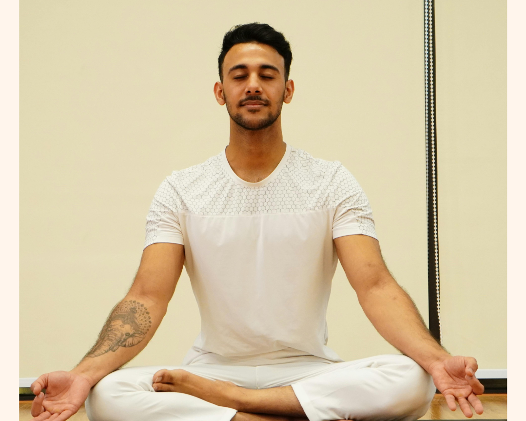 A man sitting cross-legged on a wooden floor practicing meditation with eyes closed, in a room with a light-colored wall.