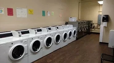 A row of front-loading washing machines in a laundromat with payment machines and a soap dispenser on the wall.