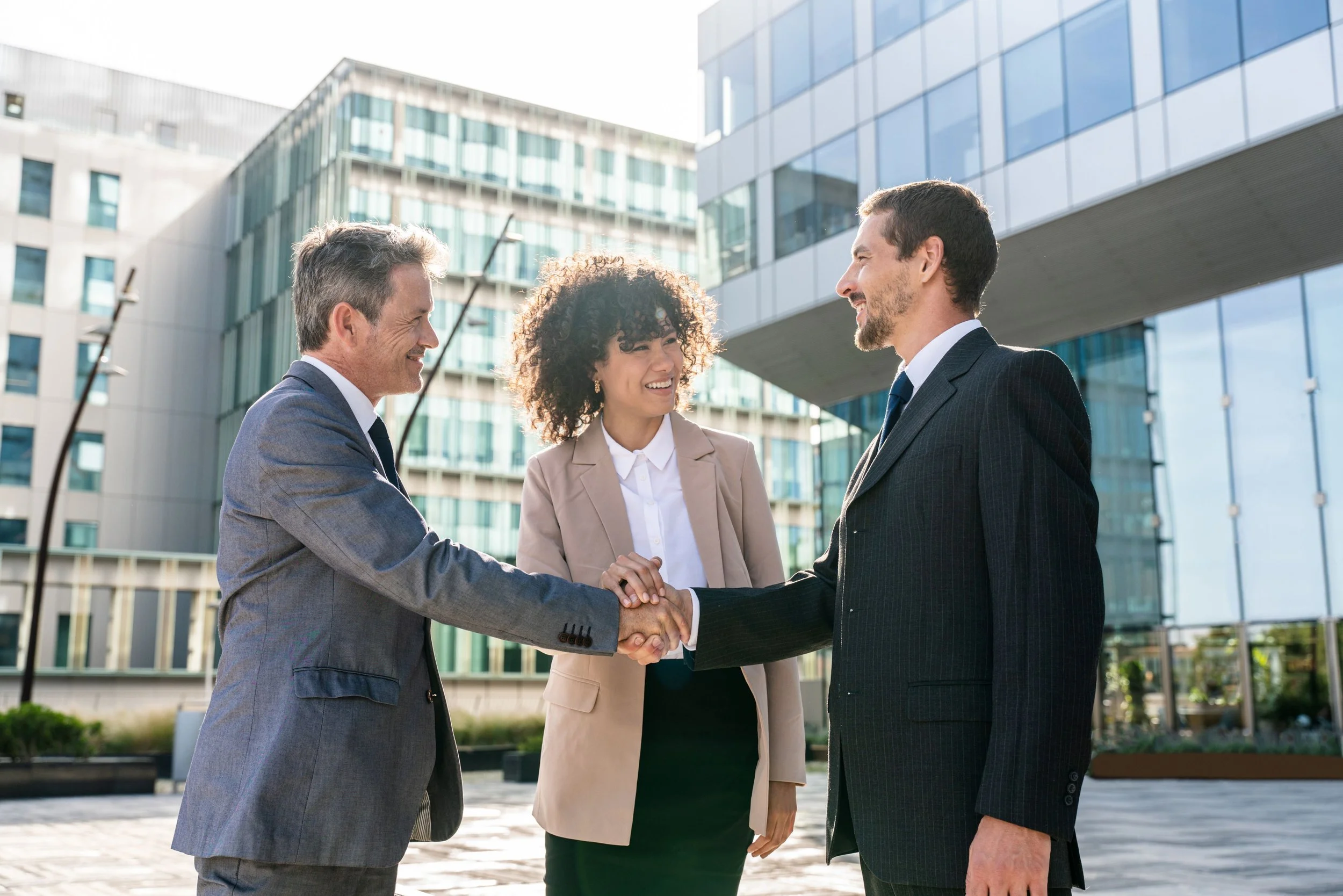 Two men and a woman shake hands outside modern office buildings, smiling and dressed in business suits.