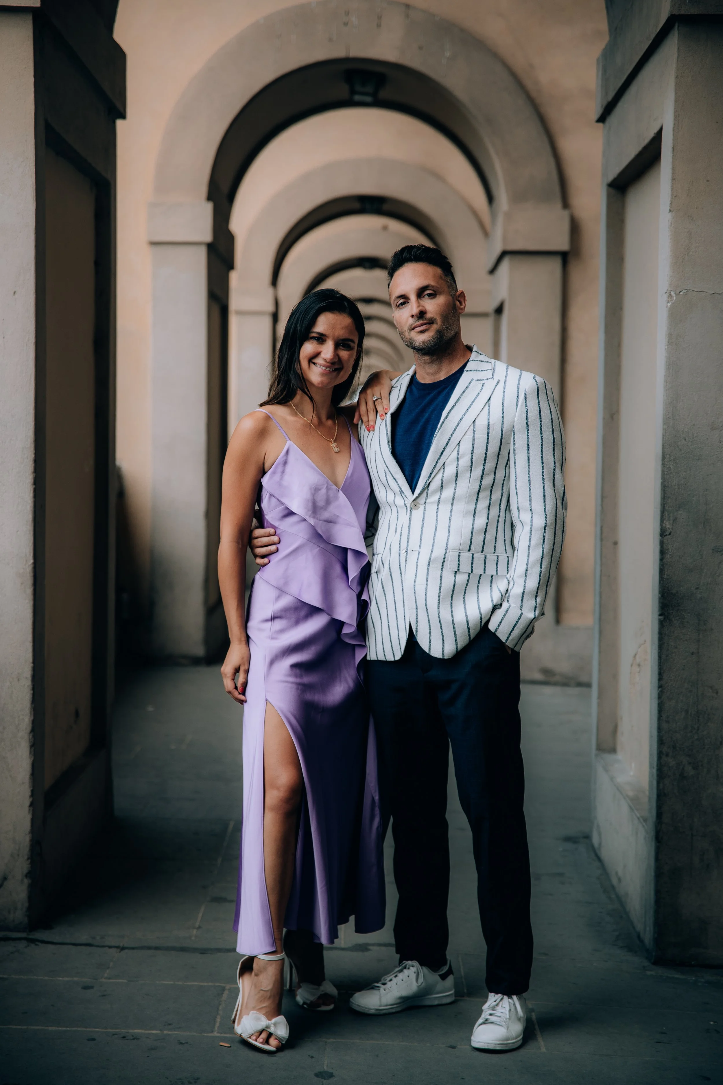 A man and woman standing together in an outdoor corridor with arched doorways, dressed in semi-formal attire.