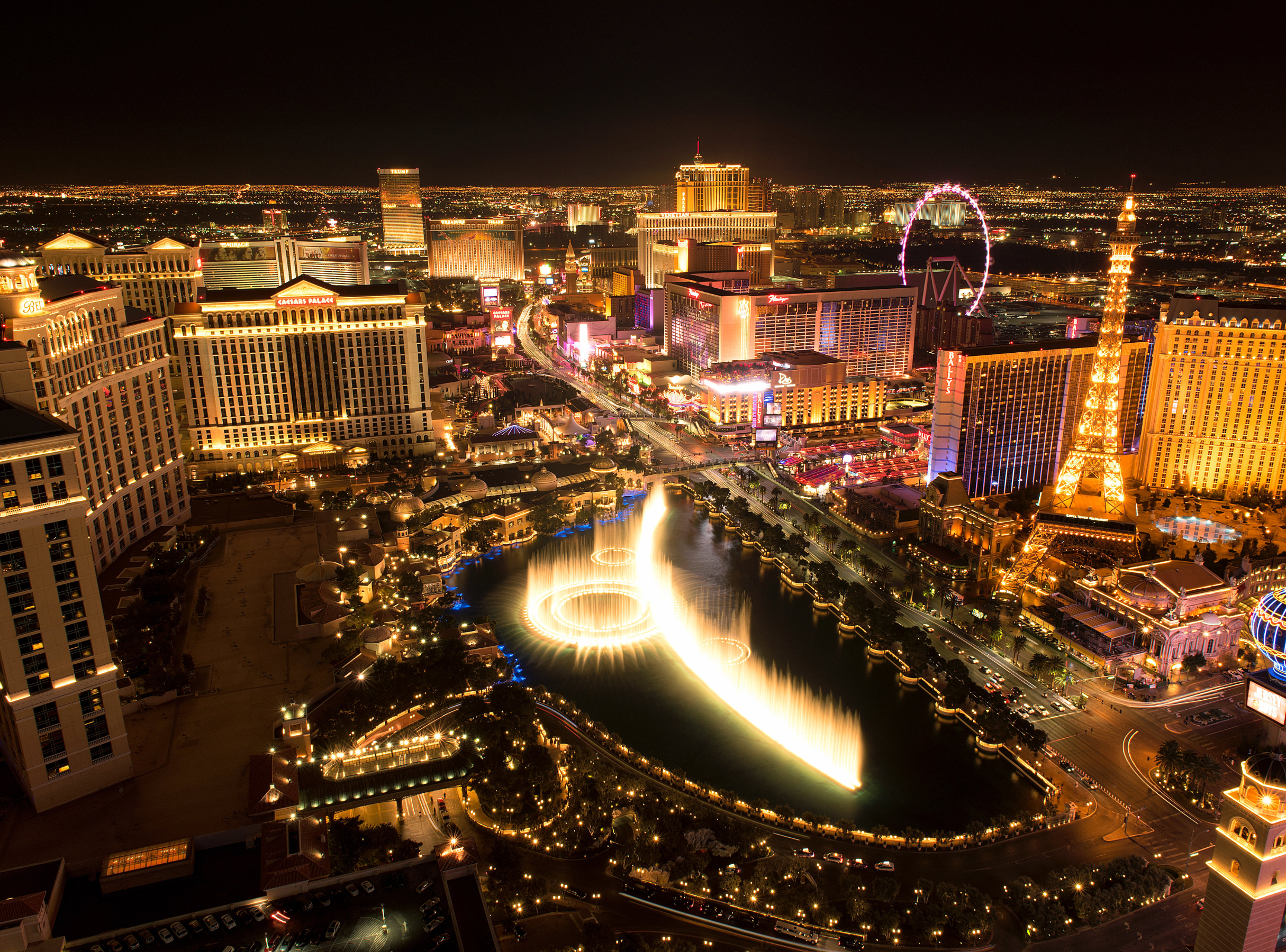 Nighttime aerial view of the Las Vegas Strip with illuminated hotels, casinos, and a large fountain display in front of a city skyline.