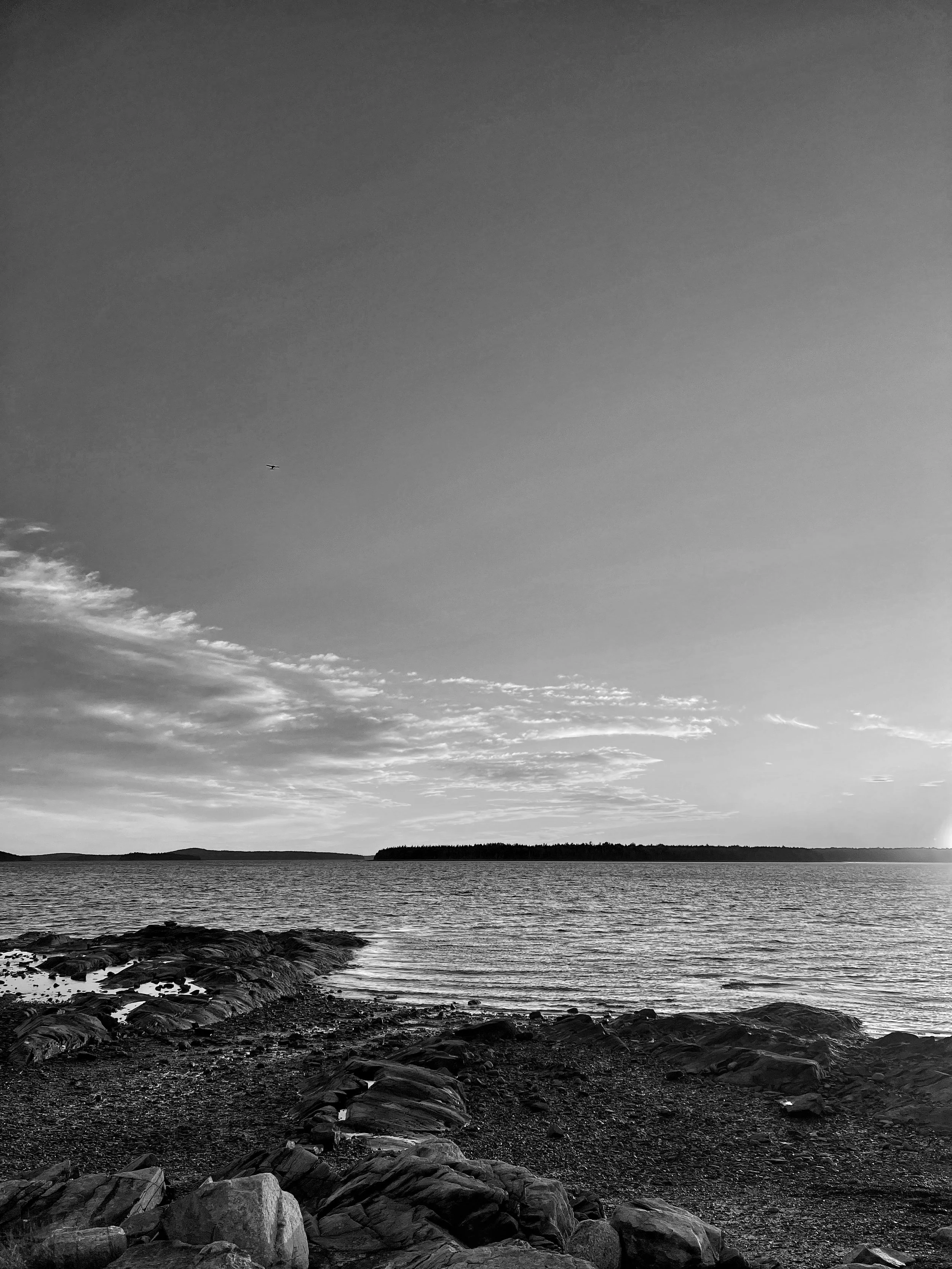 Black and white photo of a lakeshore with rocks in the foreground, water, distant land, and a sky with scattered clouds.