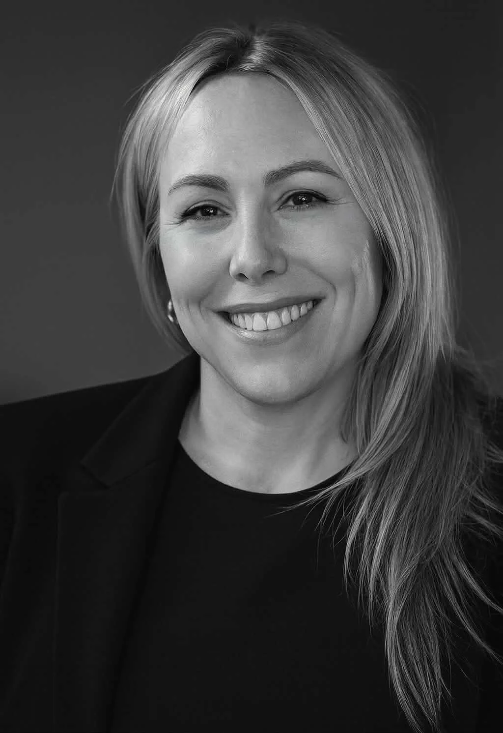Black and white portrait of a smiling woman with long, light-colored hair, wearing a dark blazer and a dark top, against a plain background. Phoenix AZ Realtor. Real Estate