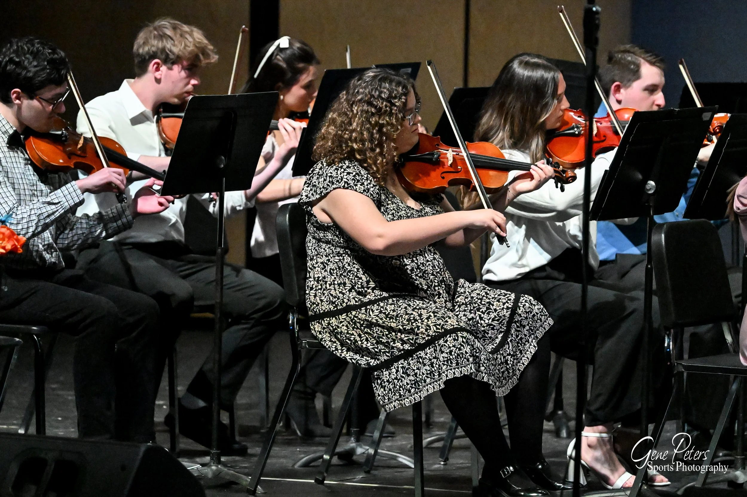Group of young musicians playing violins during a concert on stage, seated in rows with music stands in front of them.