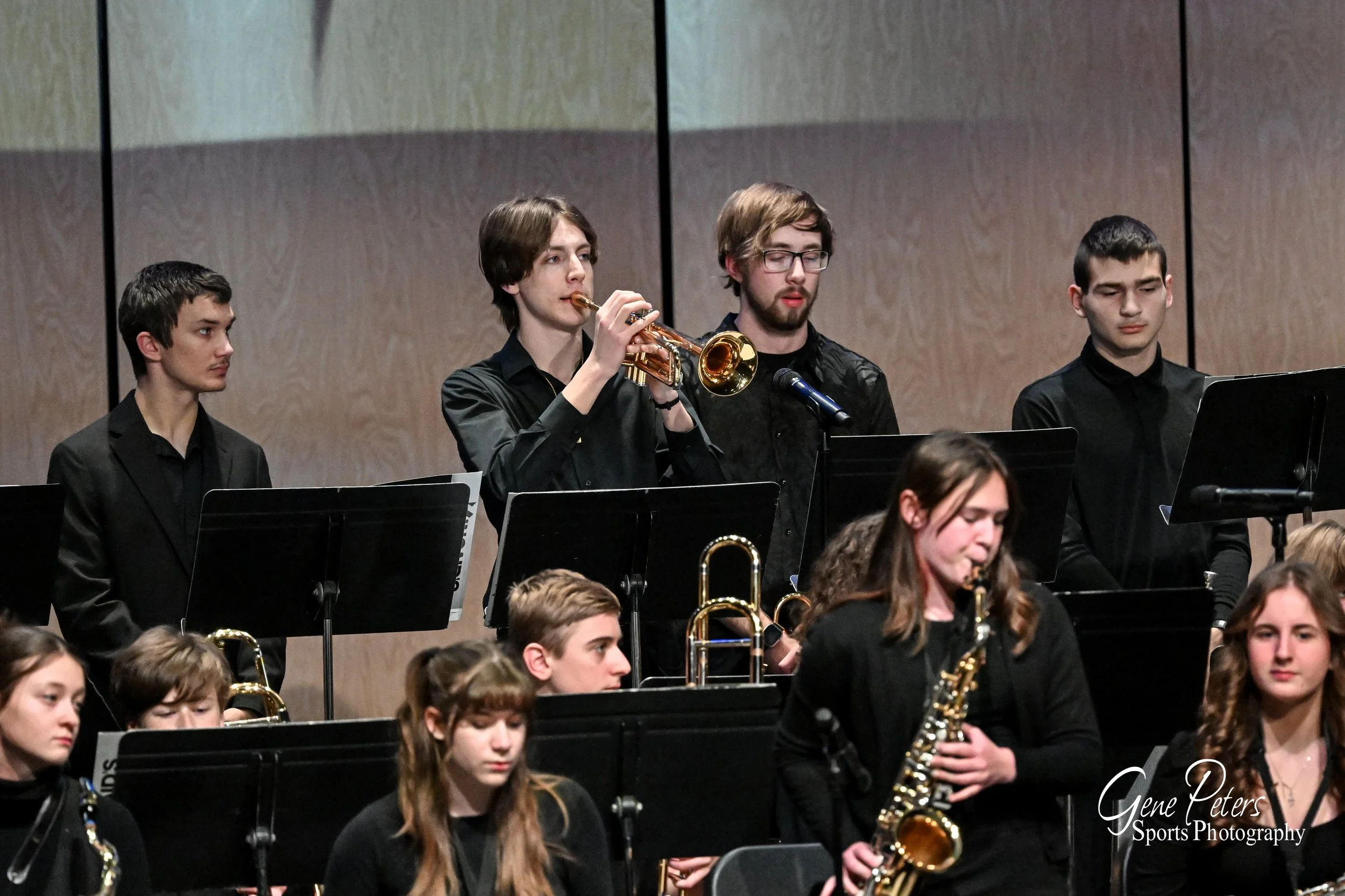 Young musicians performing in a concert hall, with some playing wind instruments such as trumpet and saxophone.