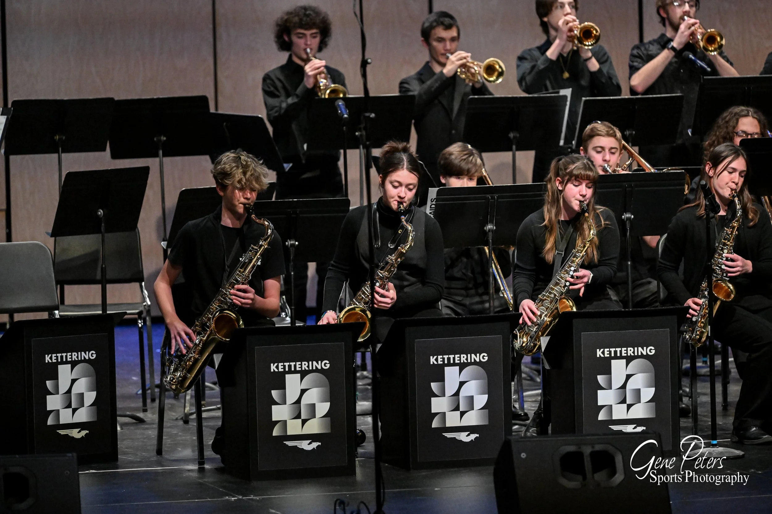 A group of young musicians playing saxophones and trumpets during a concert on stage, with music stands displaying the 'Kettering' logo.