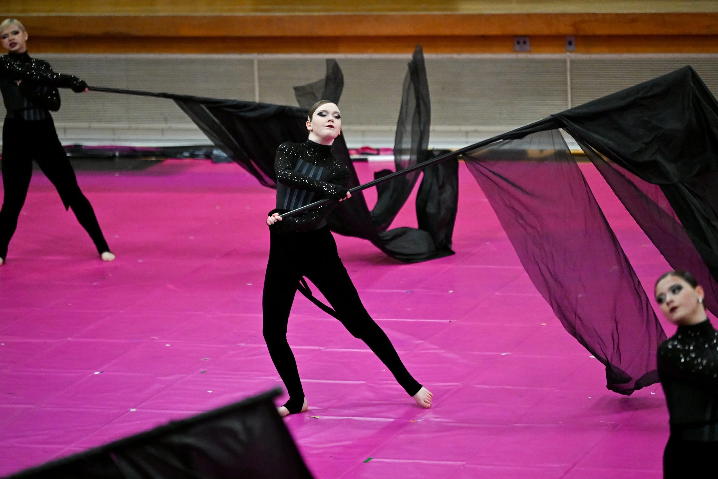 Cheerleaders performing with large black flags on a pink mat in an indoor gymnasium.