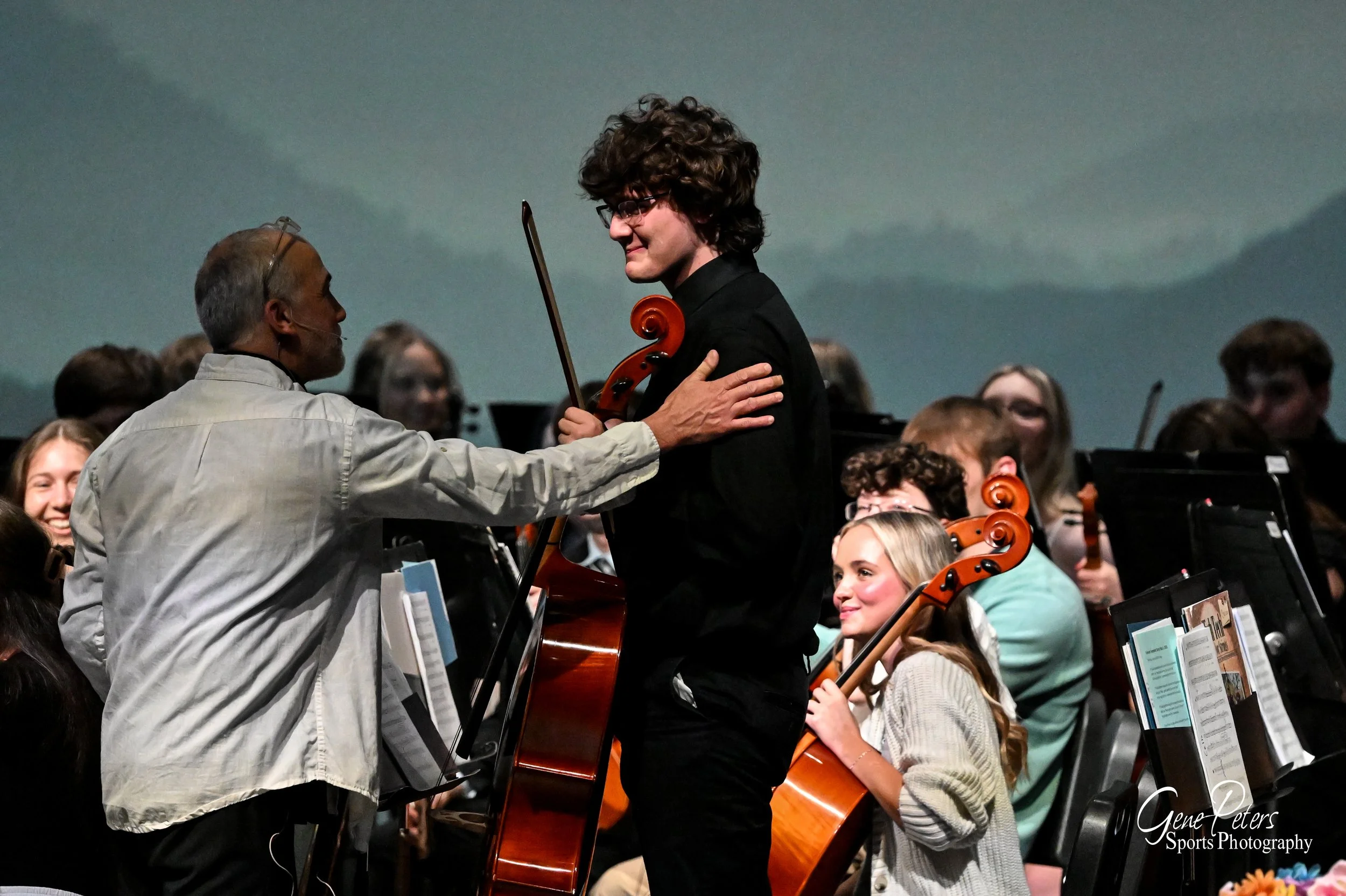 A man with glasses and a beige shirt is congratulating a young man with curly hair and glasses, who is holding a cello and a bow. They are on stage with an orchestra, including a young girl with blonde hair holding a cello, and several other musician