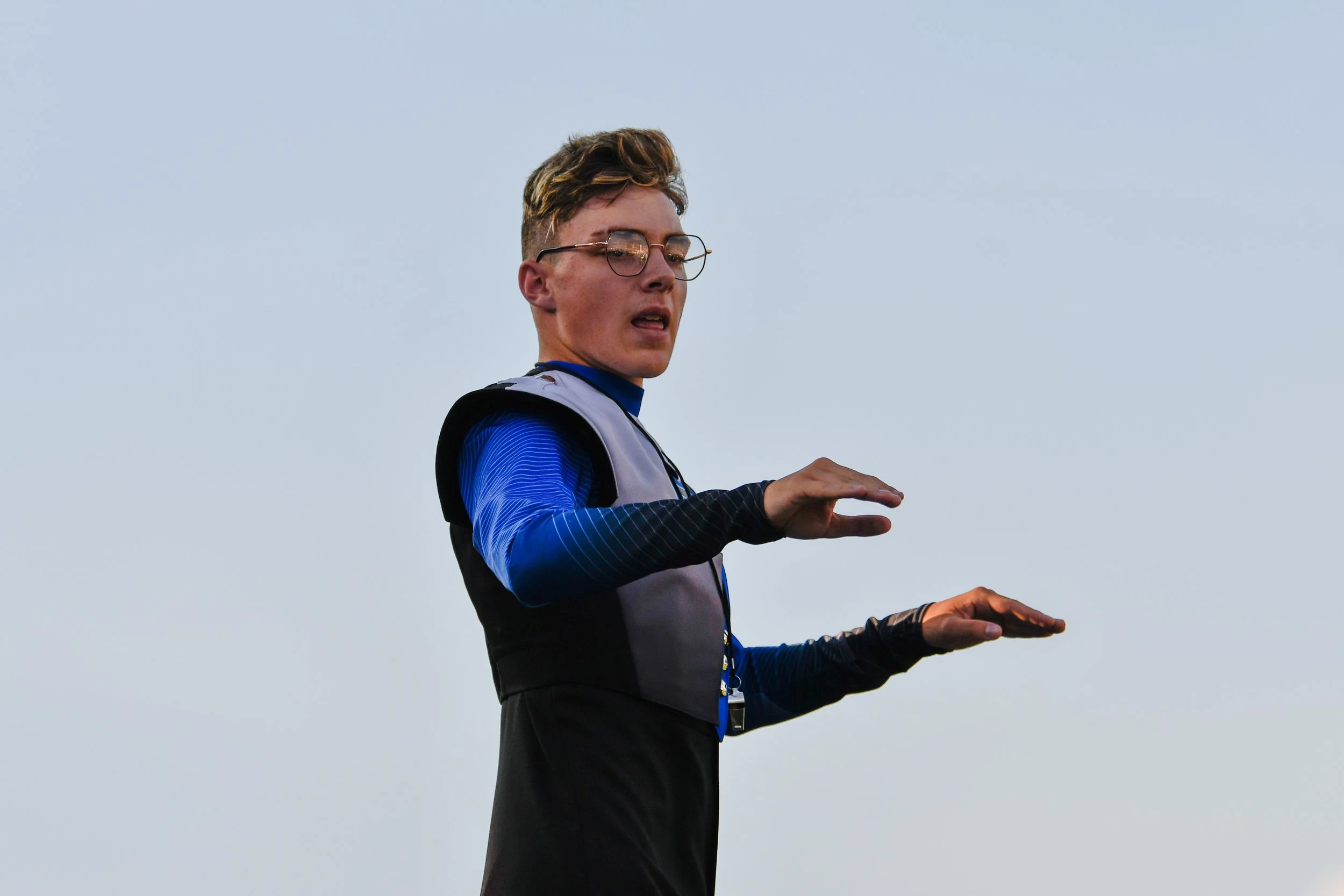 A young man with glasses, dressed in athletic clothing, is outdoors against a clear sky, holding his arms out as if signaling something or maintaining balance.