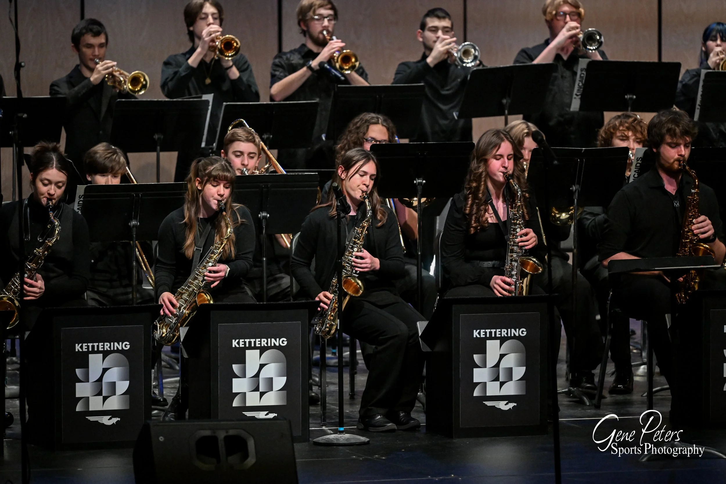 A group of musicians performing in an orchestra, playing various wind instruments such as saxophones and trumpets, with a backdrop of music stands and a stage at Kettering.