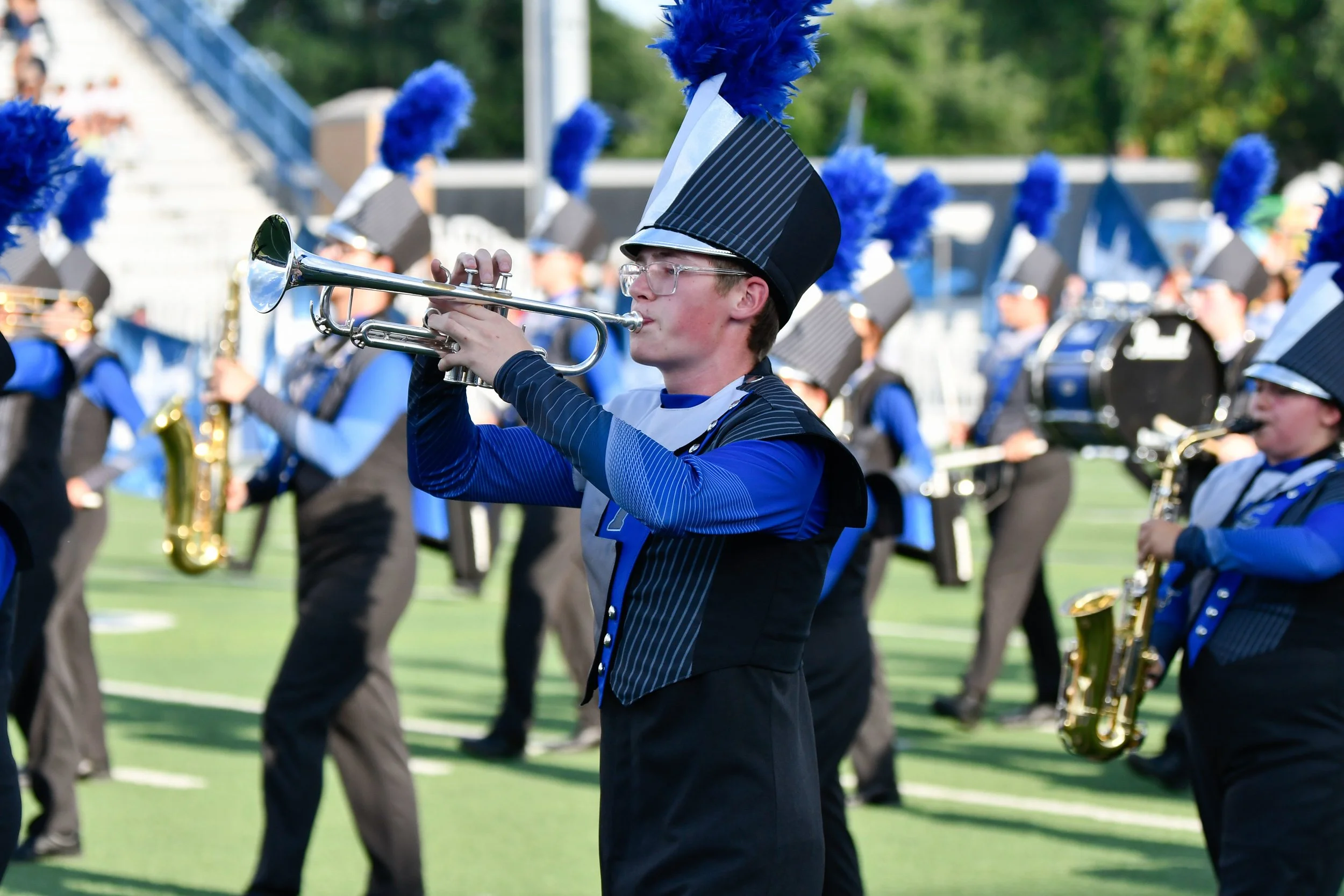 A young marching band member playing a trumpet during a parade or performance, wearing a blue and black uniform with tall black hat adorned with blue plumage, on a grassy field.