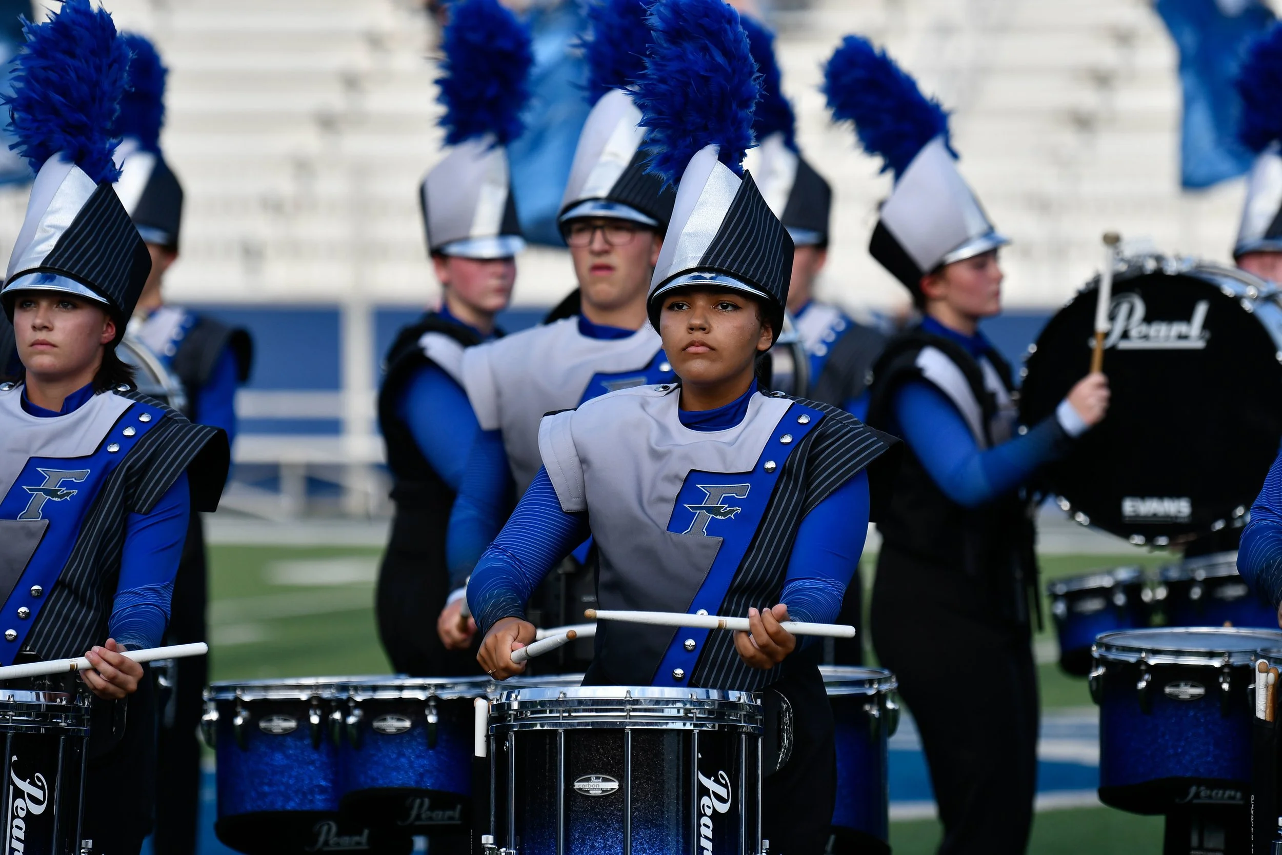 Young marching band members in blue and silver uniforms playing drums on a sports field, with a drummer holding a drumstick visible.