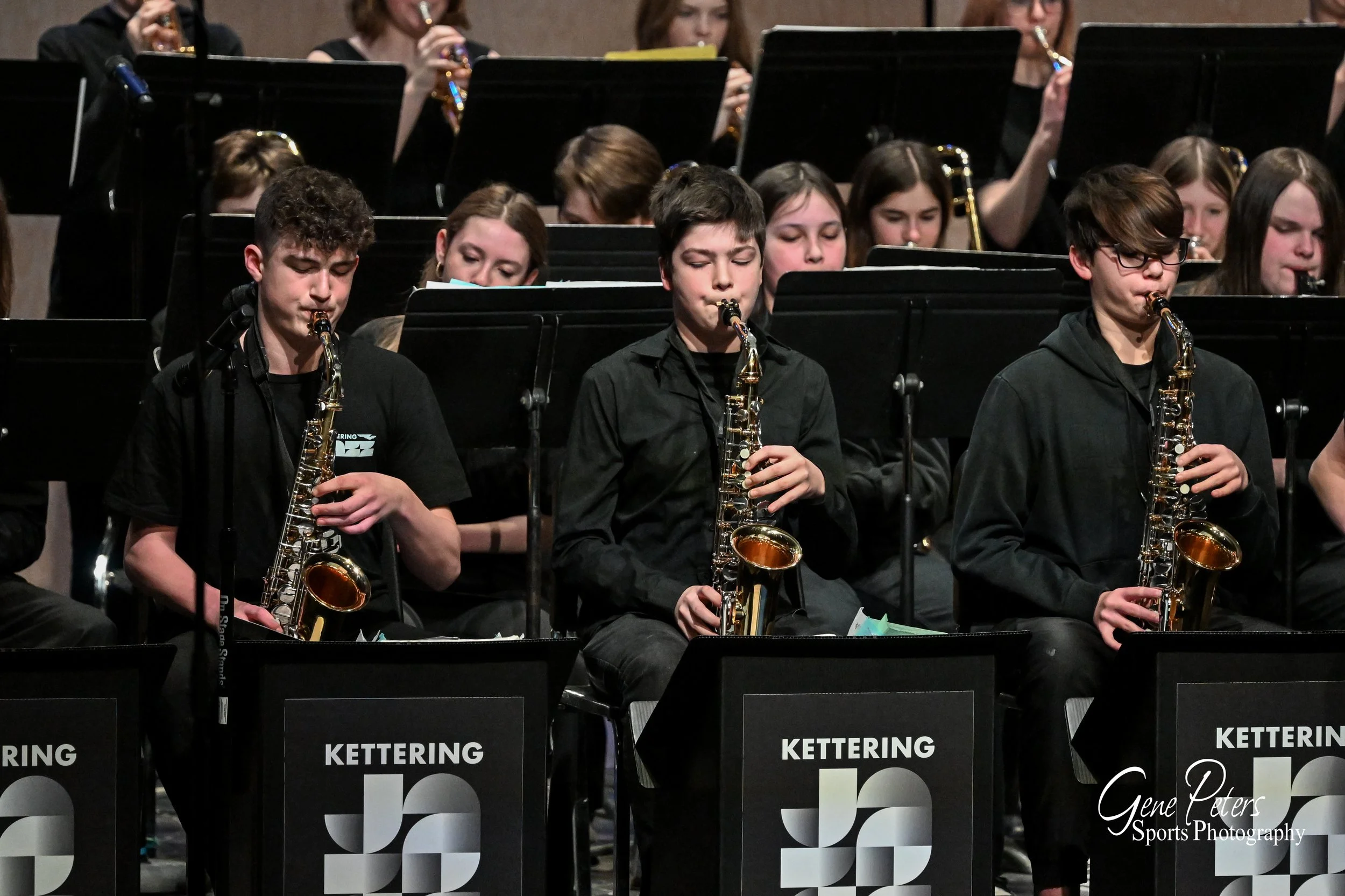 Three young musicians playing saxophones during a band concert, with other band members behind them and music stands in front labeled 'Kettering'.