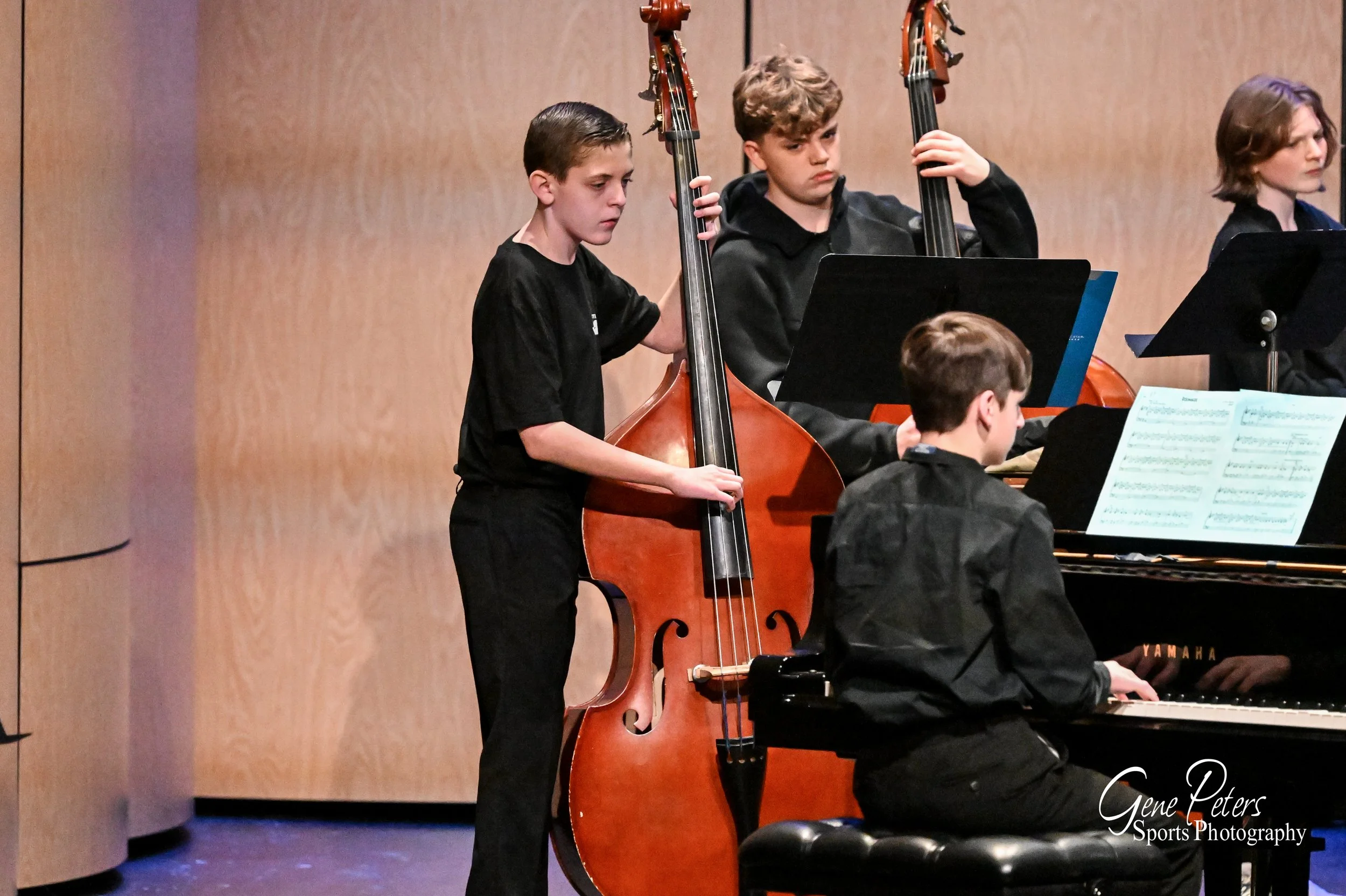 A group of boys performing in a band on stage. One boy is standing and playing a double bass, another boy is sitting at a piano, and others are standing with music sheets. The setting appears to be a concert or recital.