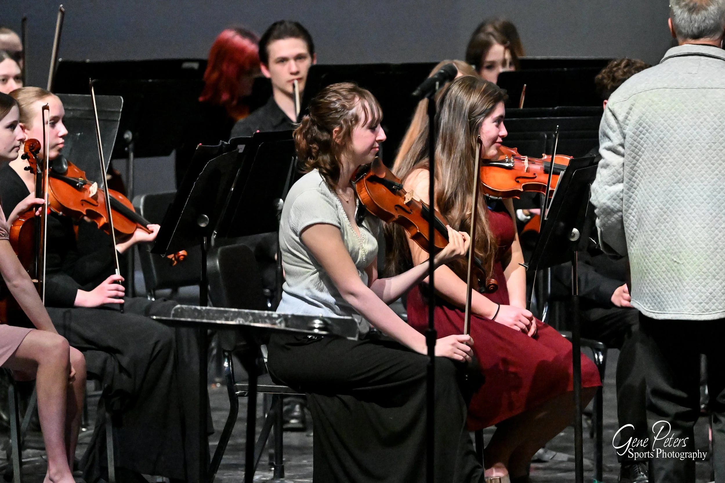 Young women playing violins in an orchestra during a performance, with a conductor standing nearby. The scene is on a stage with music stands and other musicians visible in the background.