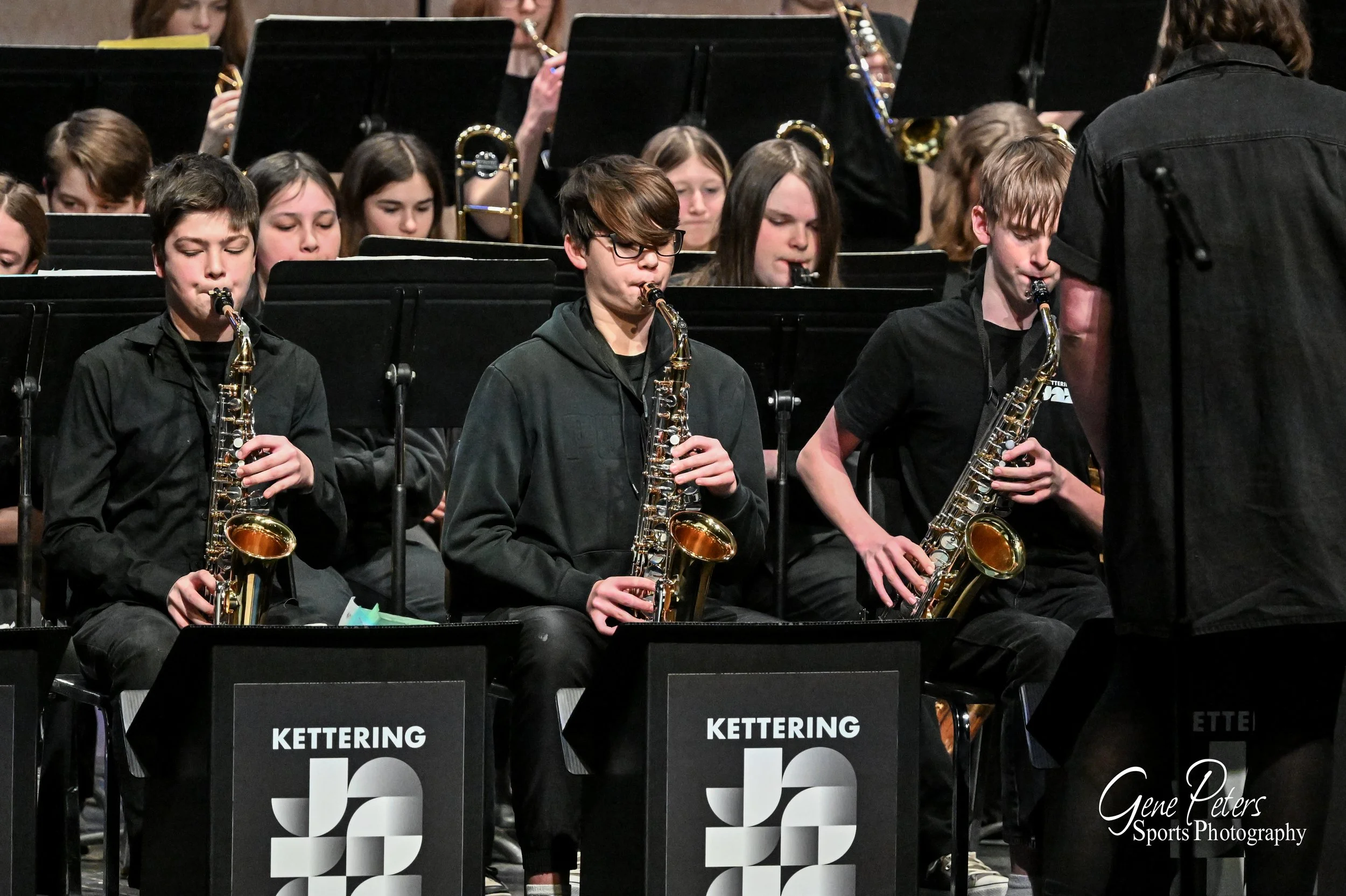 Young musicians playing saxophones in a concert, dressed in black, with music stands bearing the Kettering name, conducted by an instructor, with other band members in the background.