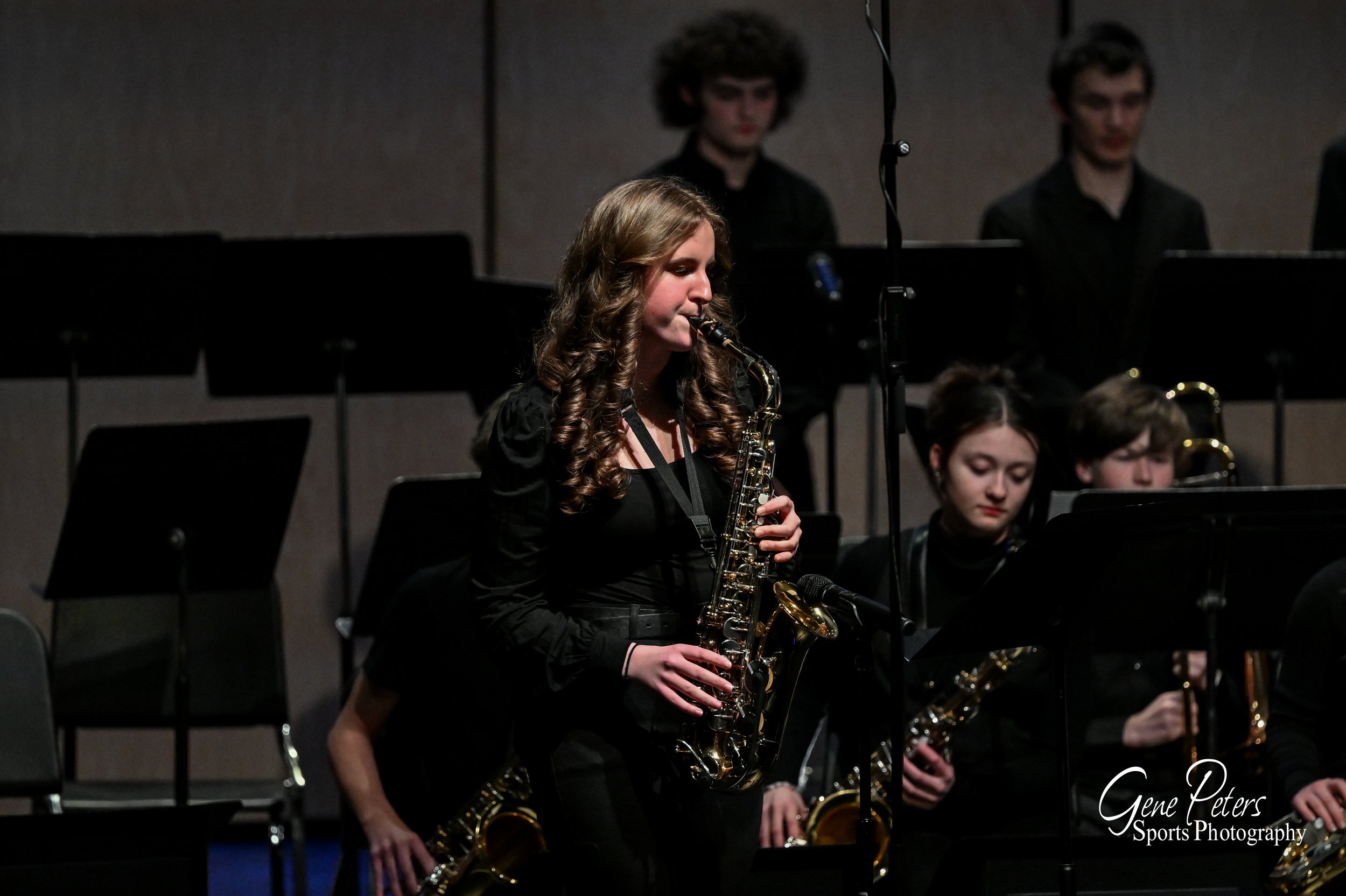 A young woman with long curly hair playing a saxophone during a concert, surrounded by other musicians on stage.